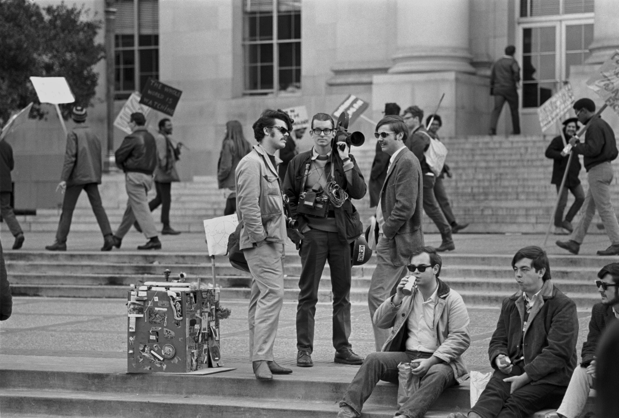 A group of men stand and sit on outdoor steps; some hold cameras, others are talking or drinking. People walk by with protest signs in the background near a large building with tall windows.
