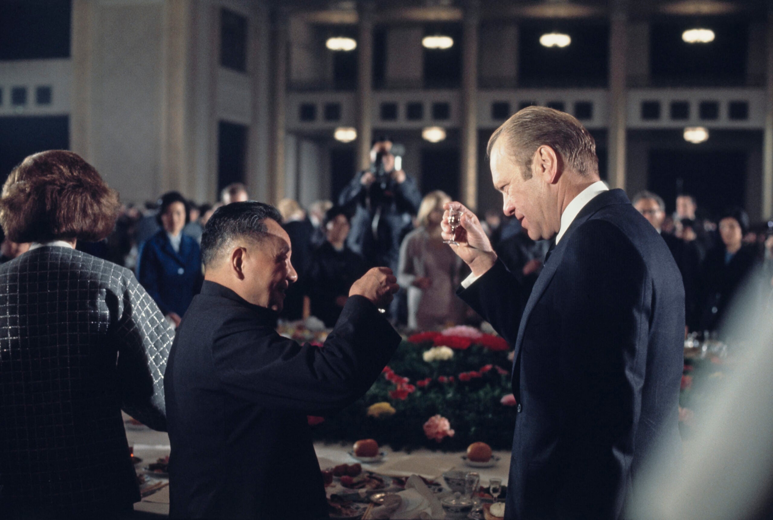 Two men in suits raise glasses in a toast at a formal banquet, surrounded by other guests and photographers in a large, elegant room with high ceilings and chandeliers.