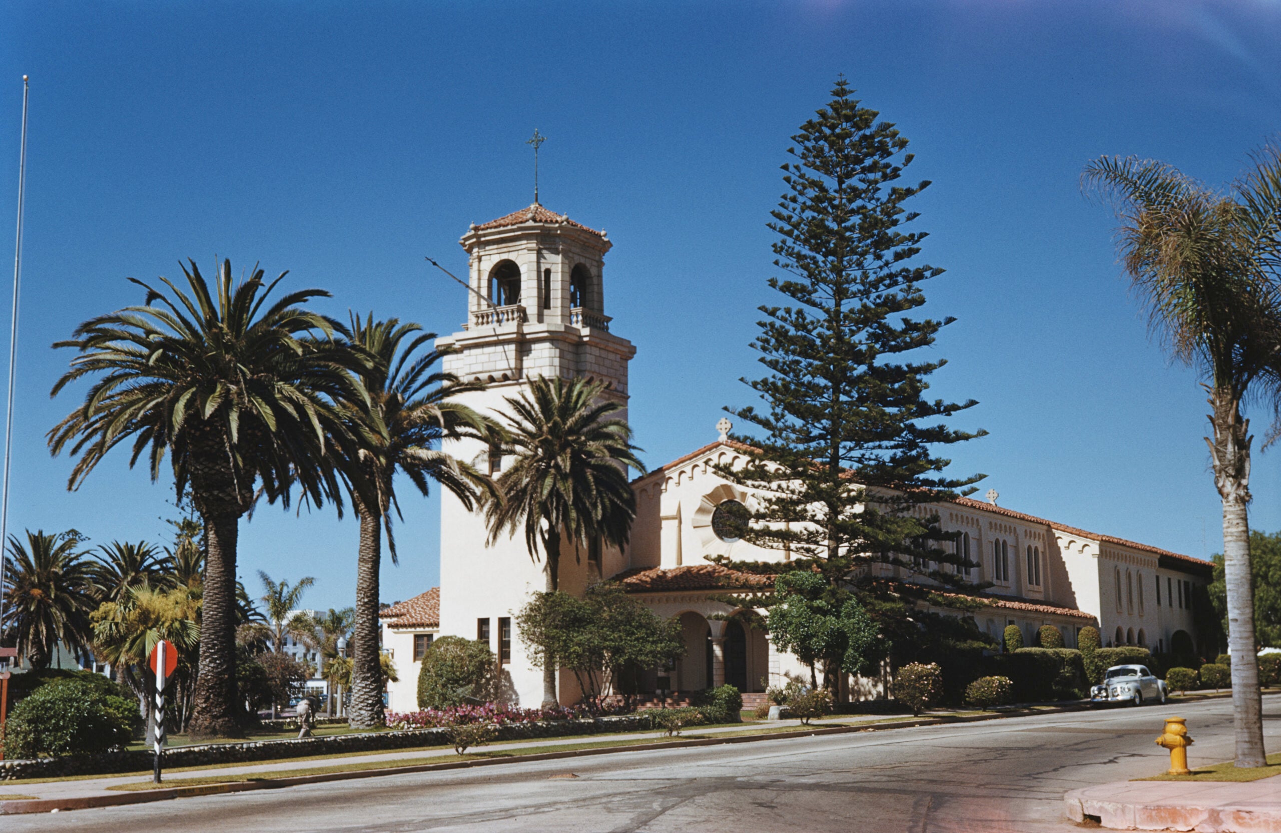 A Spanish-style building with a bell tower, palm trees, and a tall evergreen is shown under a clear blue sky. A yellow fire hydrant sits on the corner of the street in the foreground.