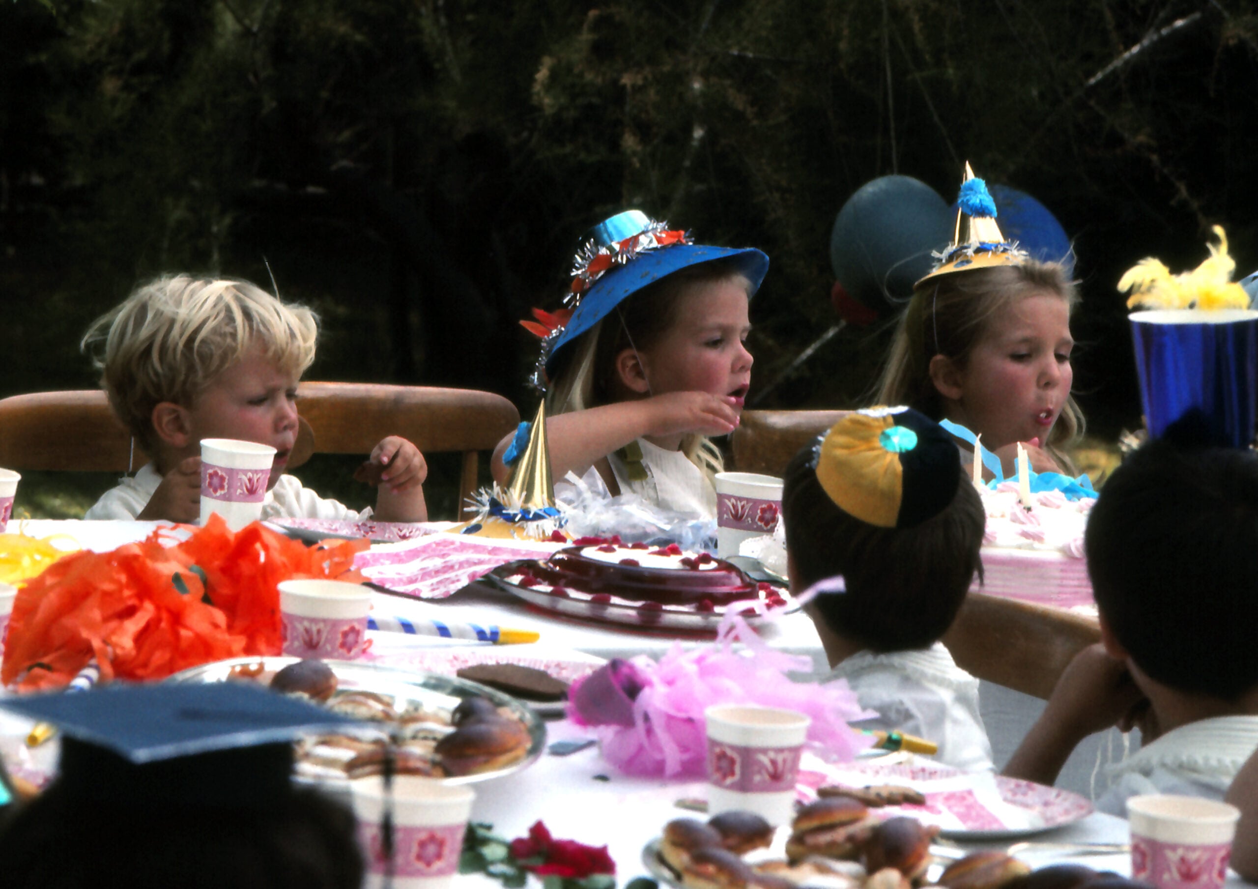 Children wearing party hats sit around a table outdoors, eating cake and snacks at a festive birthday party. Balloons and colorful decorations are on the table.