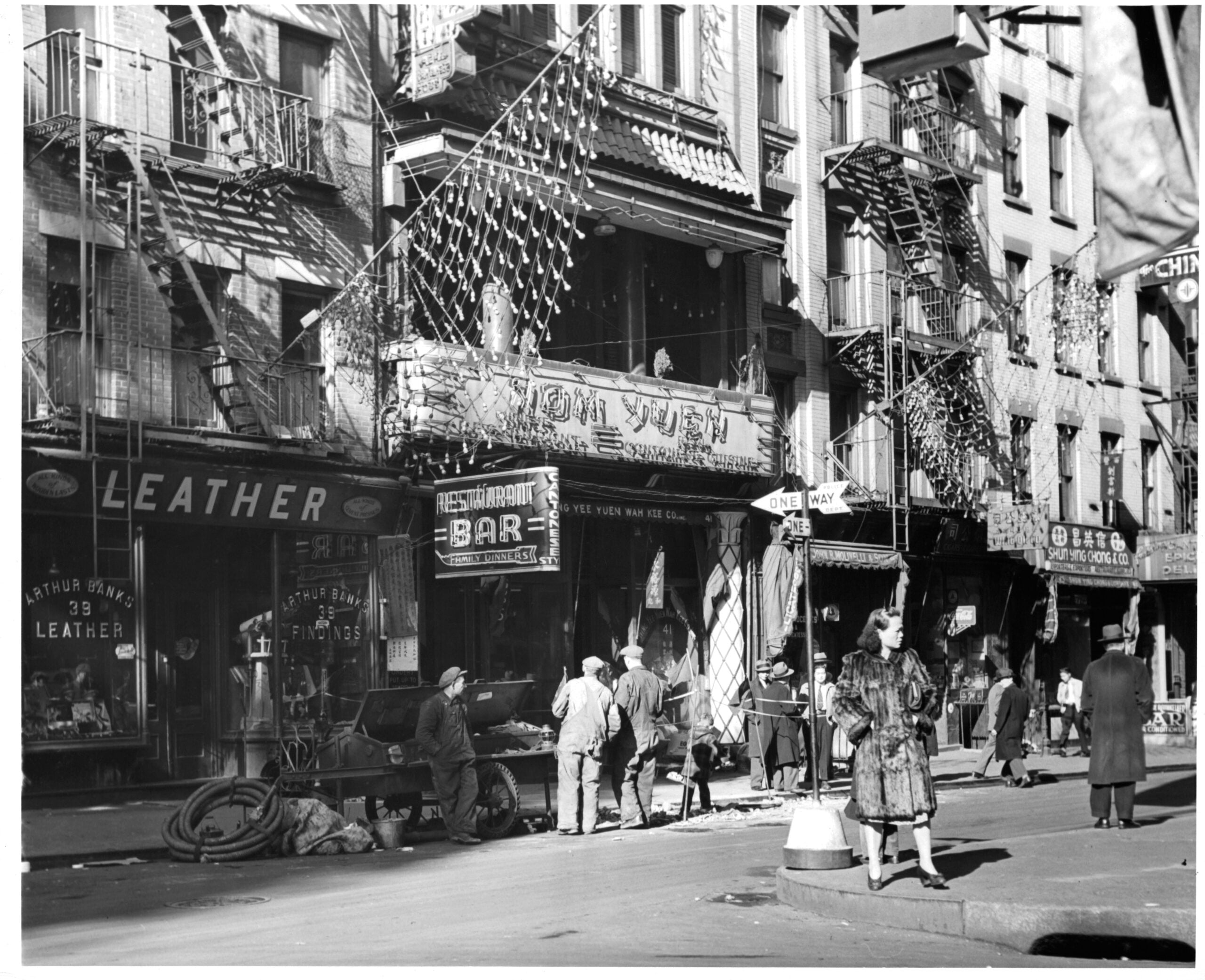 Black-and-white photo of a busy city street with people walking, a restaurant bar, shops, fire escapes on buildings, and festive string decorations overhead. The scene appears to be from the mid-20th century.
