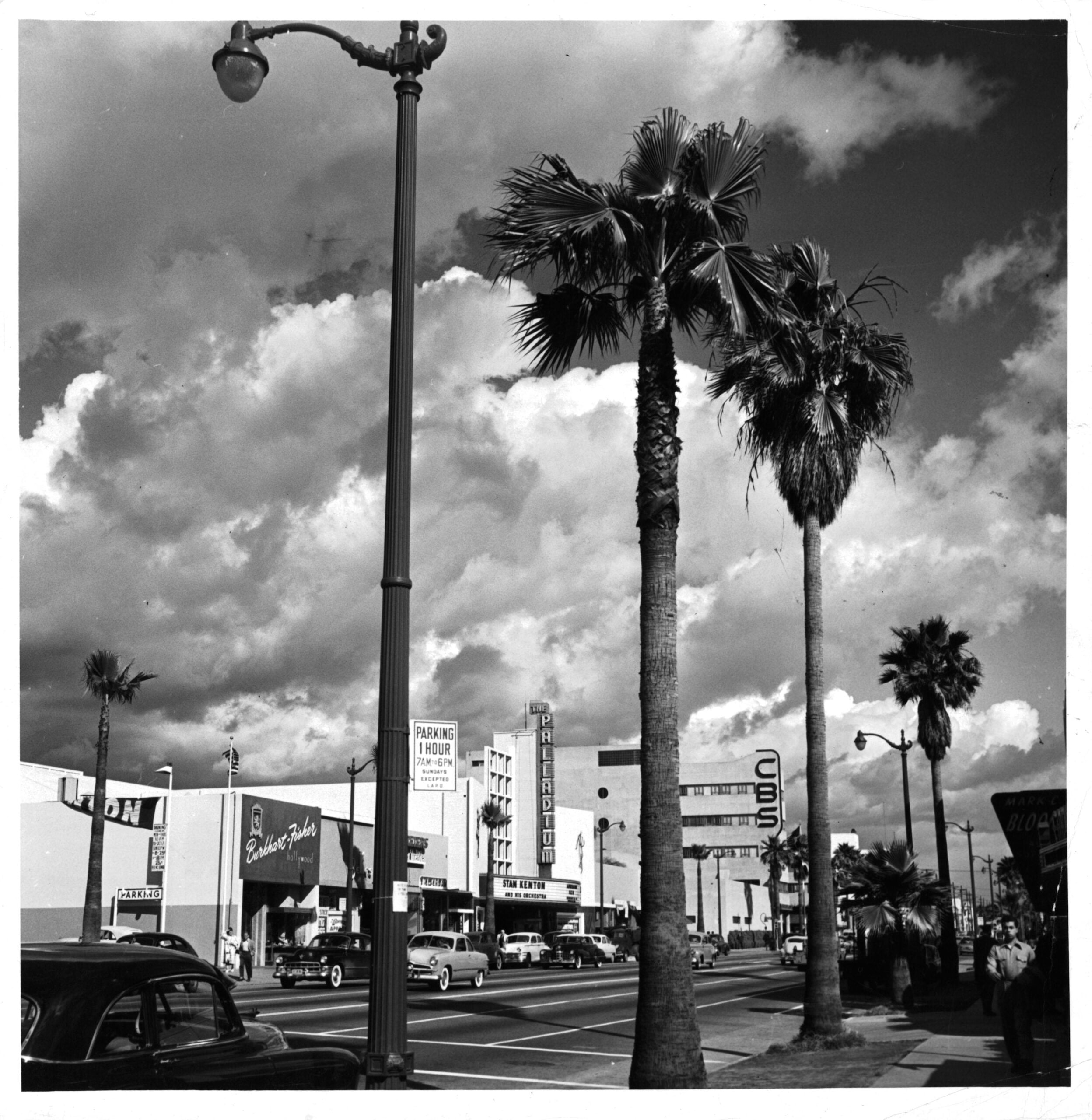 Black-and-white photo of a city street lined with palm trees, vintage cars, and buildings. The CBS studio sign is visible among other storefronts. The sky is dramatic with large, billowing clouds.