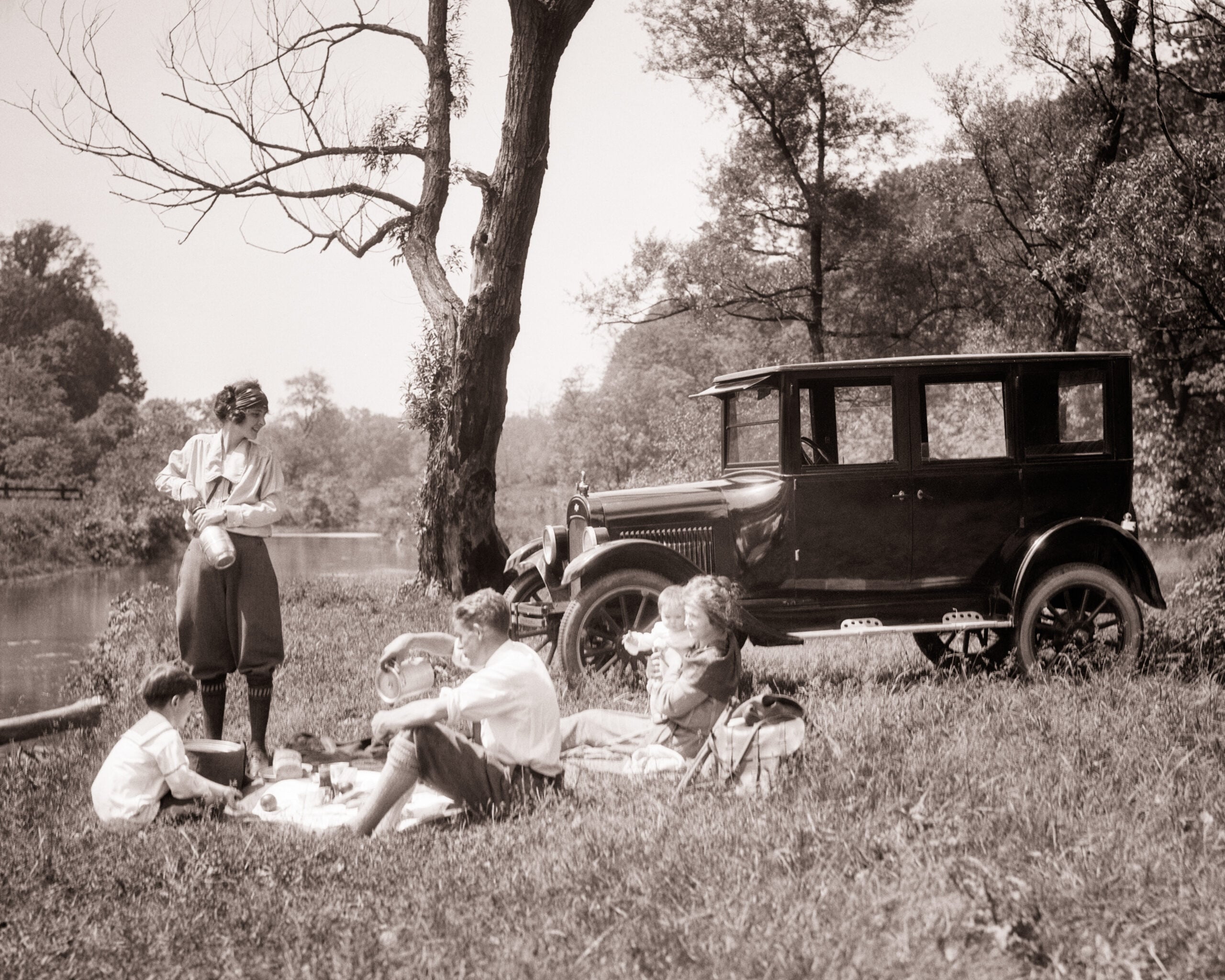 A vintage car is parked by a river as a family of four enjoys a picnic on the grass. Trees surround them under a clear sky, and the scene appears to be from the early 20th century.