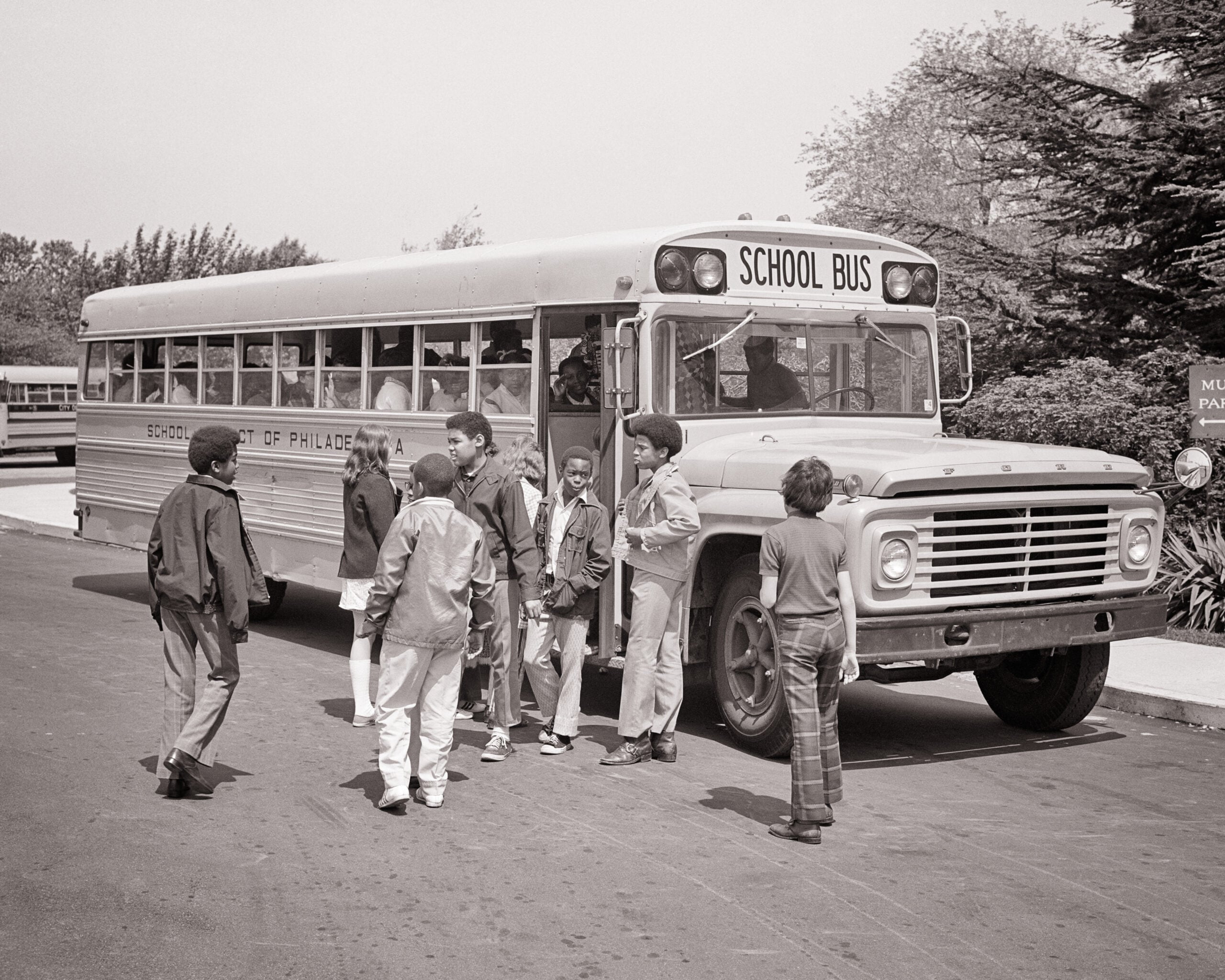 A group of children stands near the open door of a school bus, talking and preparing to board. The bus is parked on a paved area with trees in the background.