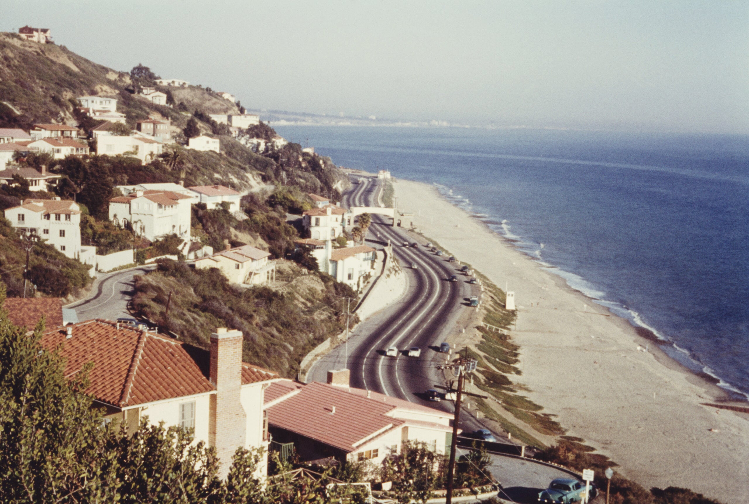 A coastal road runs alongside the beach, bordered by houses on a hill. The ocean is calm, and a few cars are visible on the road. The scene suggests a peaceful, sunny day.