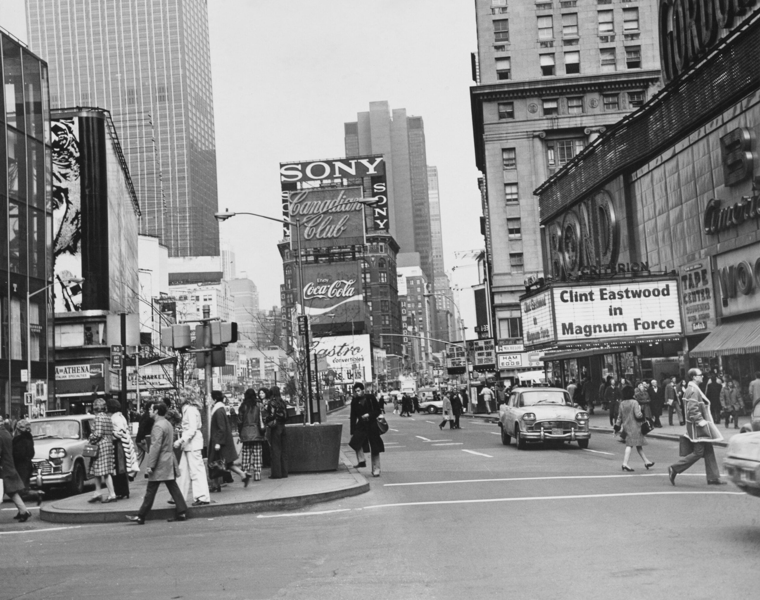 Black-and-white photo of a busy city intersection with people walking, vintage cars, and tall buildings. Billboards and a “Clint Eastwood in Magnum Force” movie marquee are visible.
