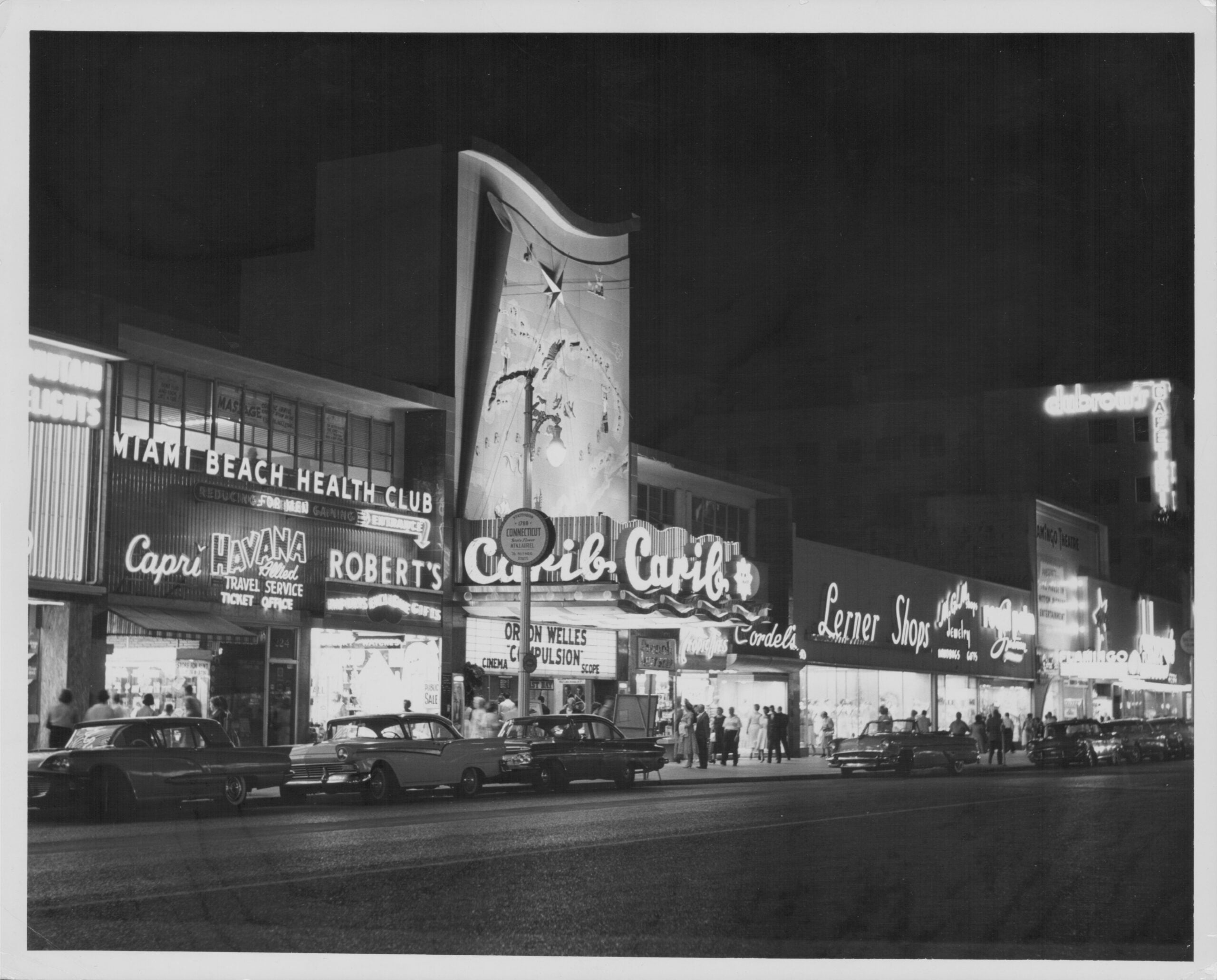 Black and white photo of a city street at night with neon signs for clubs, shops, and restaurants. Vintage cars are parked along the street, and people walk on the sidewalk.