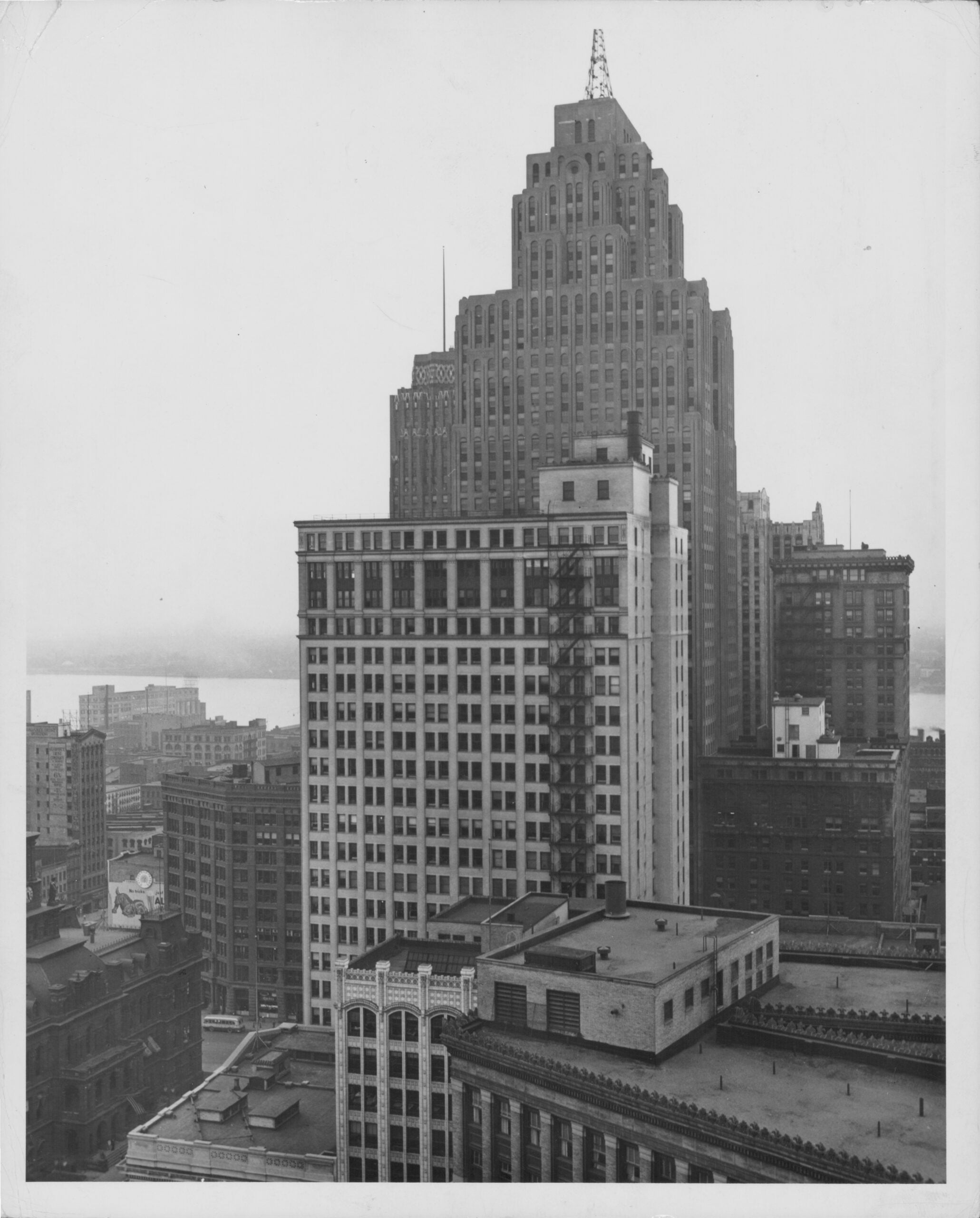 Black-and-white photo of a city skyline with tall, tiered skyscrapers, mid-rise buildings, and rooftops in the foreground; water and more buildings can be seen in the background.
