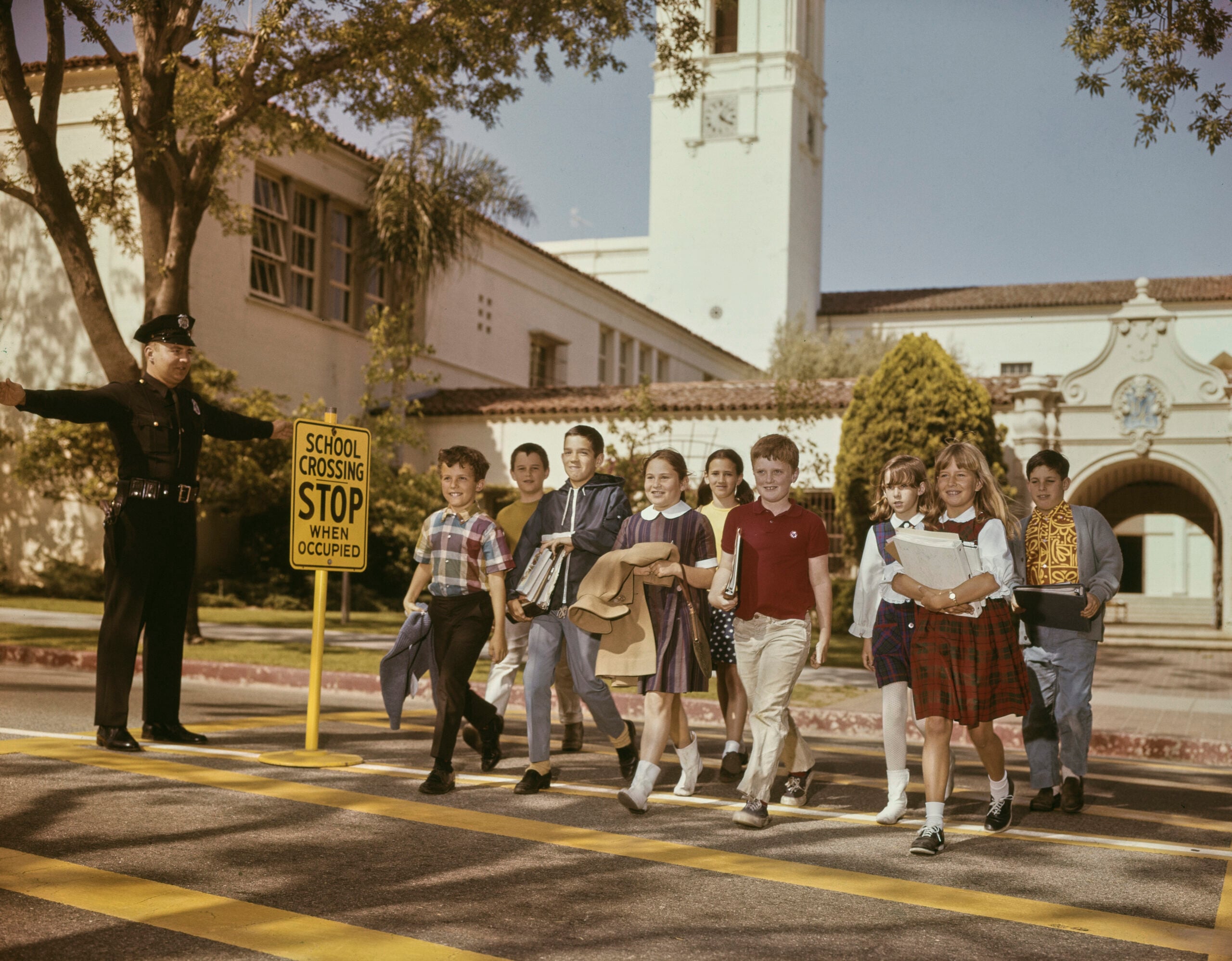 A police officer stops traffic as a group of schoolchildren cross a street at a crosswalk in front of a school building on a sunny day, capturing a classic moment from school life in the 1960s and 70s.