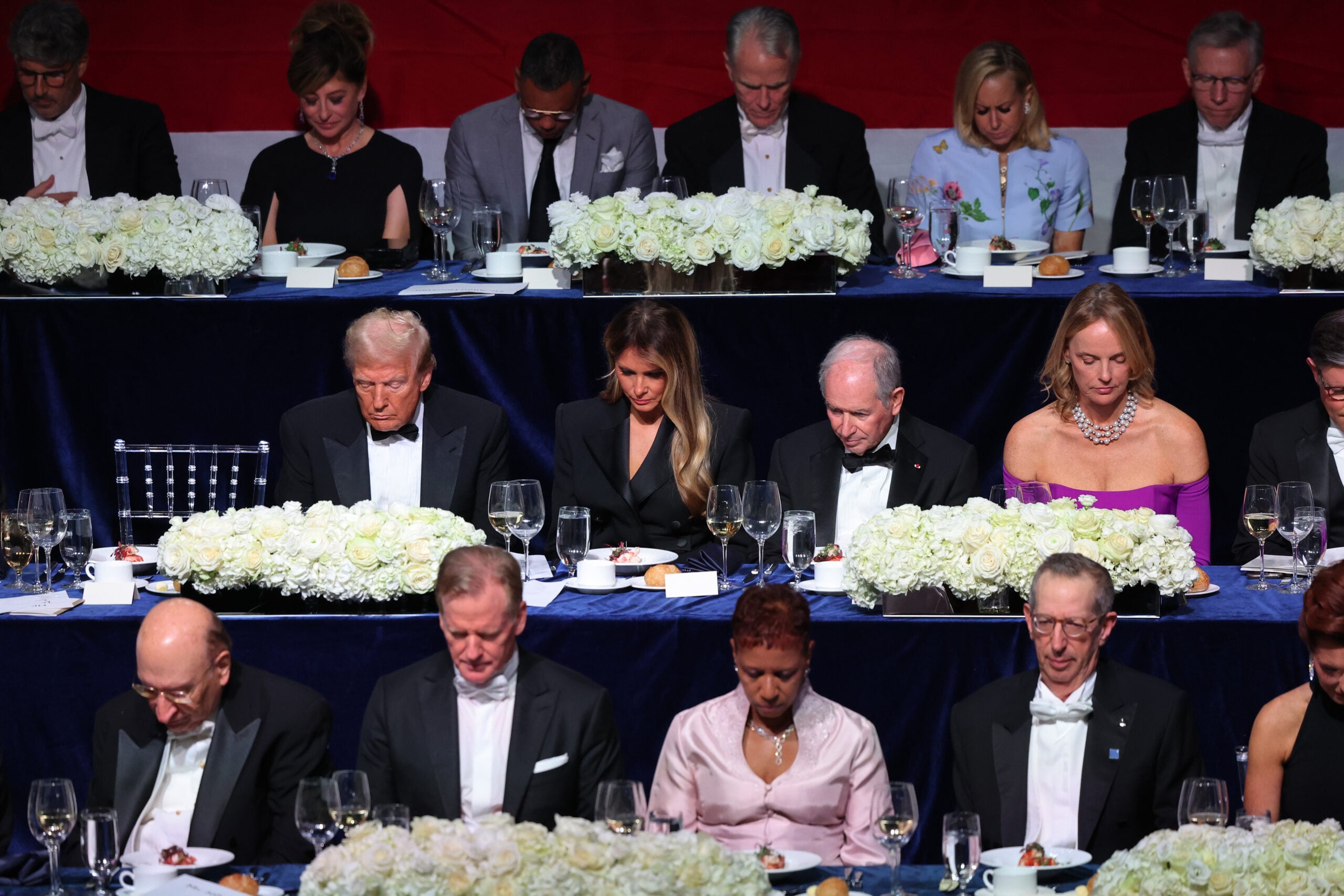 A group of elegantly dressed people sit at long banquet tables, heads bowed in apparent prayer or reflection. The tables are decorated with white flowers, and plates and glasses are set for a formal dinner.