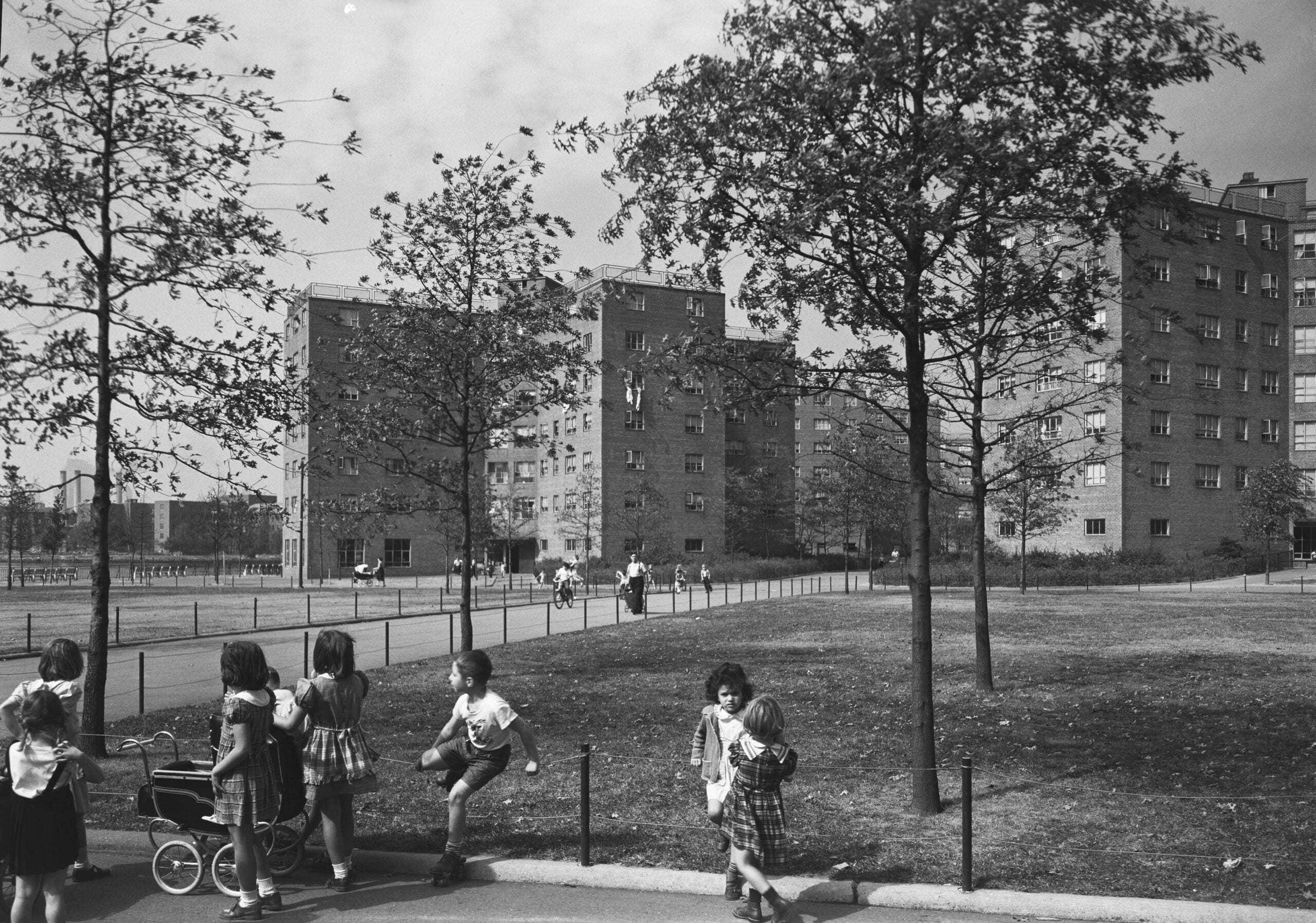 Childhood in the 50s and 60s. Children play in a grassy park area with trees, near apartment buildings in the background. Some kids are running, while others stand or push a toy stroller. The scene appears to be from a past decade.