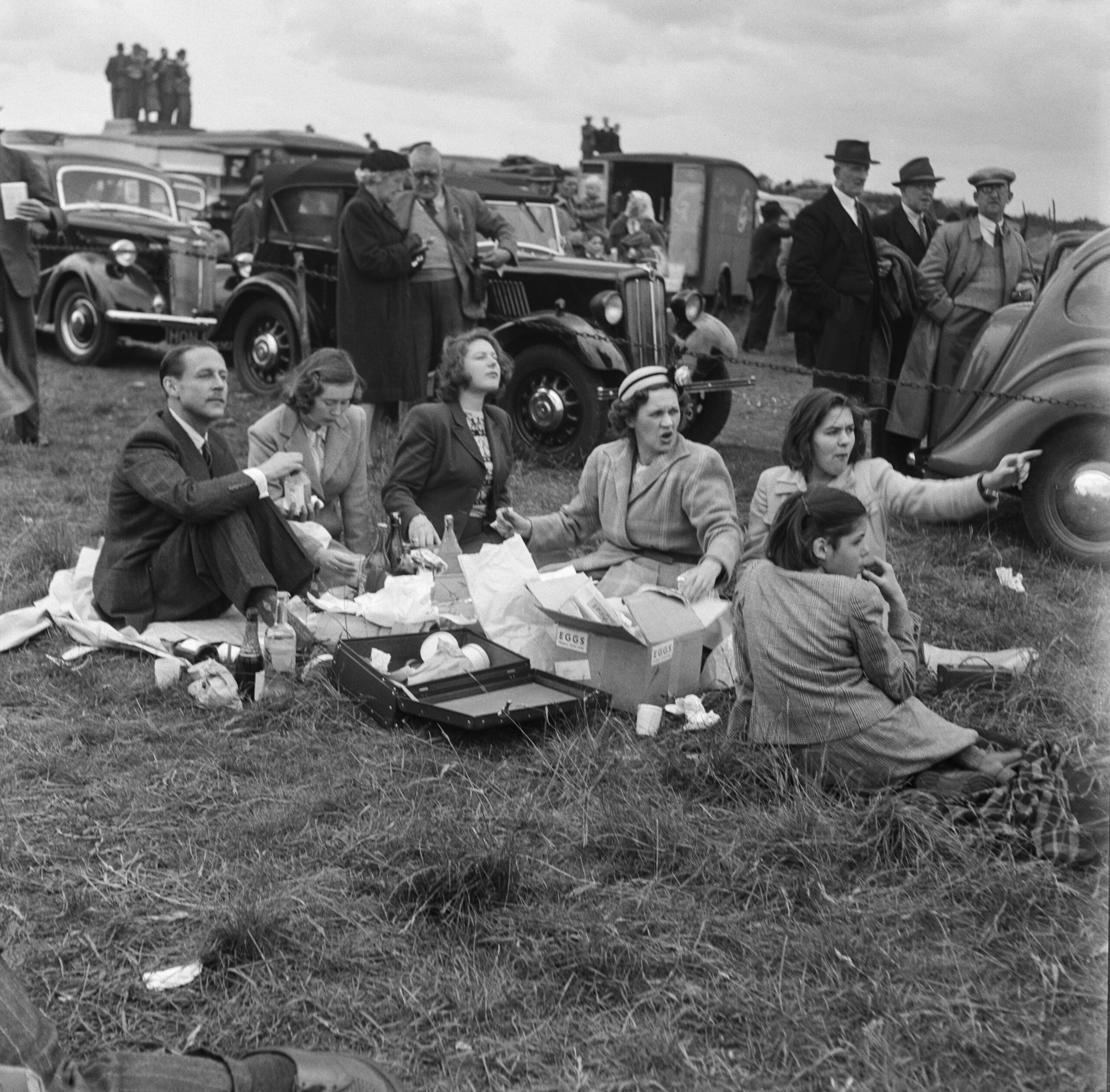 A group of people sit on picnic blankets eating and talking in a grassy field, with vintage cars and more people standing in the background under a cloudy sky.
