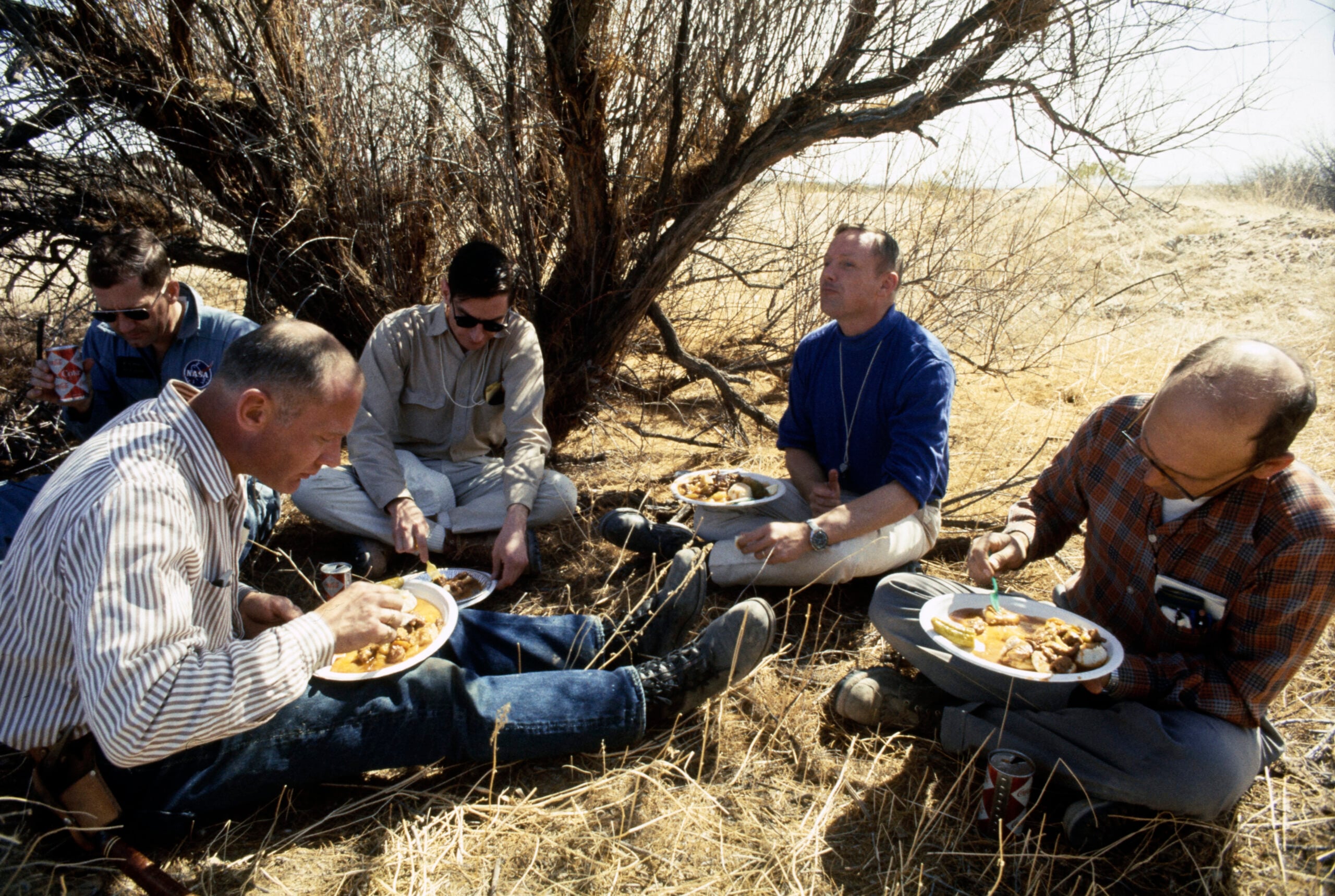 Five men sit on dry grass under a tree, eating food from plates. They appear relaxed, enjoying a meal outdoors. One man has a can of soda next to him. The scene looks sunny and arid, with leafless branches overhead.