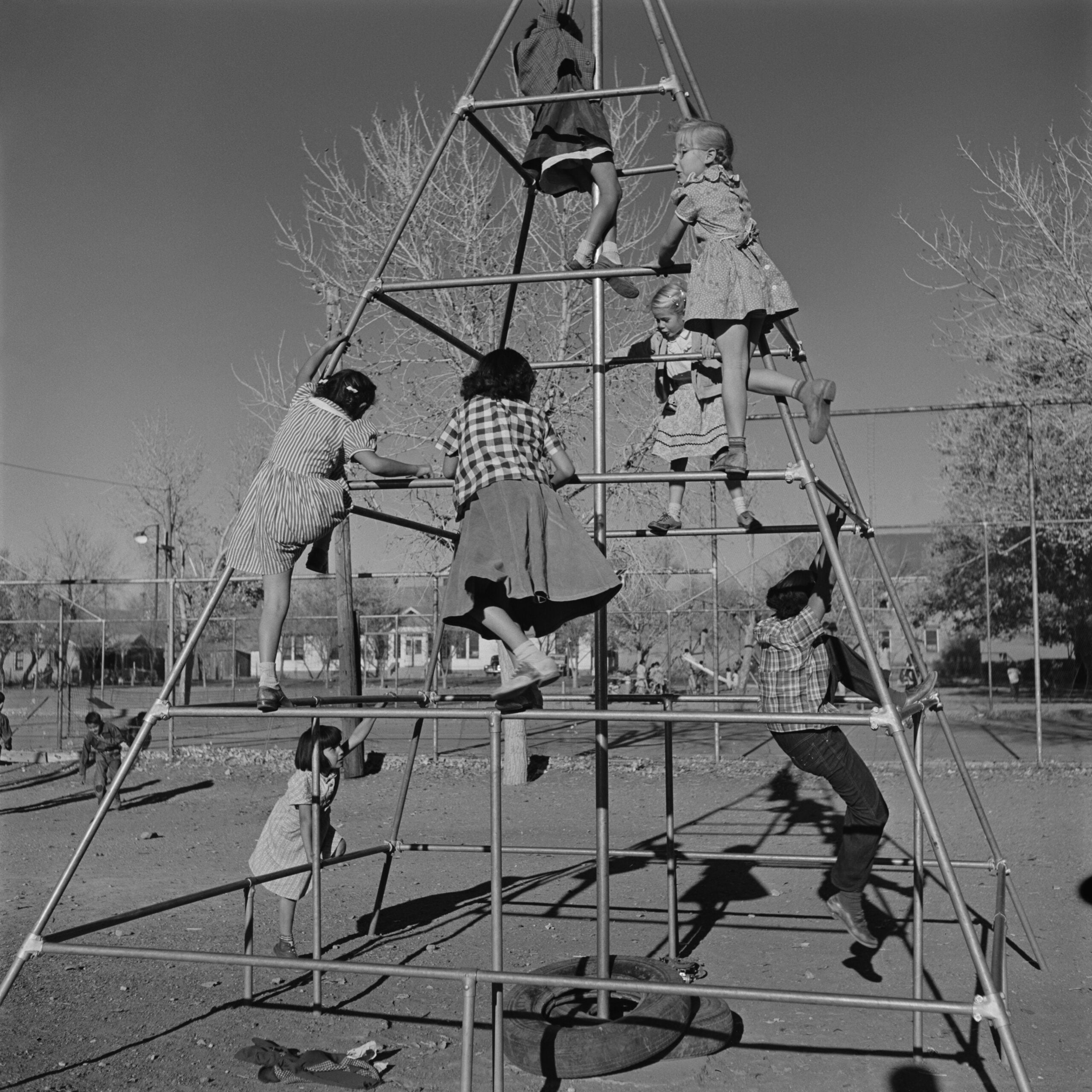 Children climb a metal jungle gym shaped like a pyramid in a playground. The scene is outdoors, with leafless trees and buildings in the background. The kids wear patterned clothes and dresses. It is a sunny day.