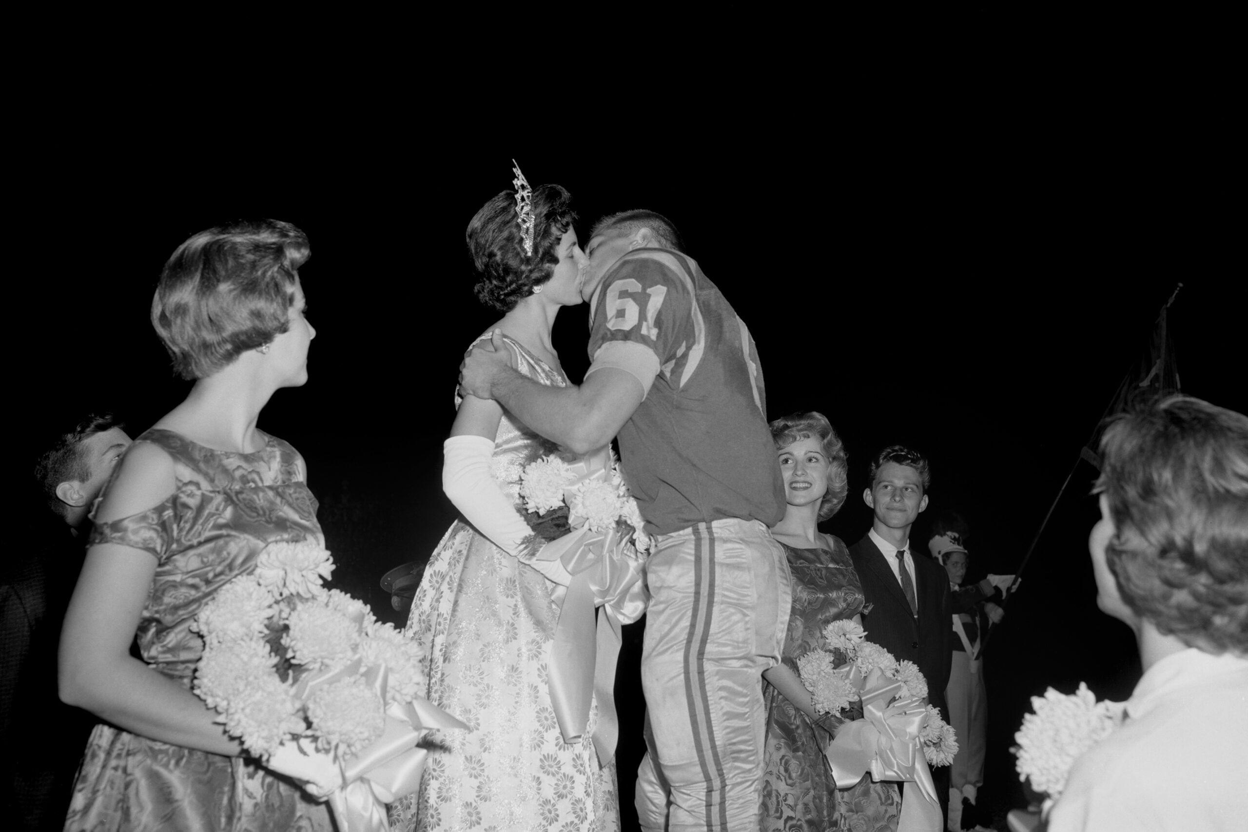 A football player in uniform kisses a woman wearing a crown and gown, surrounded by other women holding flowers at night, capturing the quintessential spirit of school life in the 1960s and 70s—possibly during a classic homecoming event.