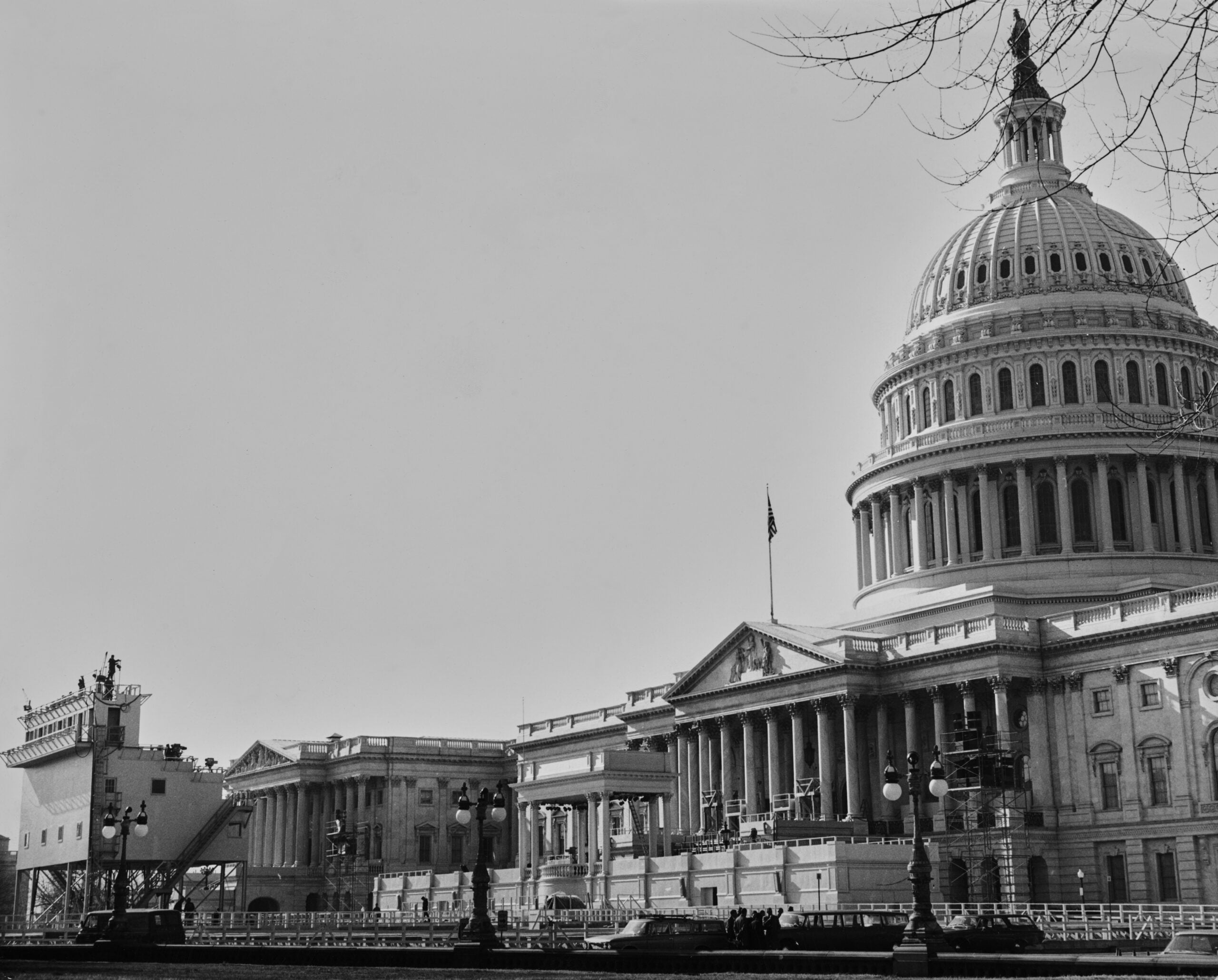 Black-and-white photo of the U.S. Capitol building with a temporary stage set up outside, and bare tree branches visible in the upper right corner.