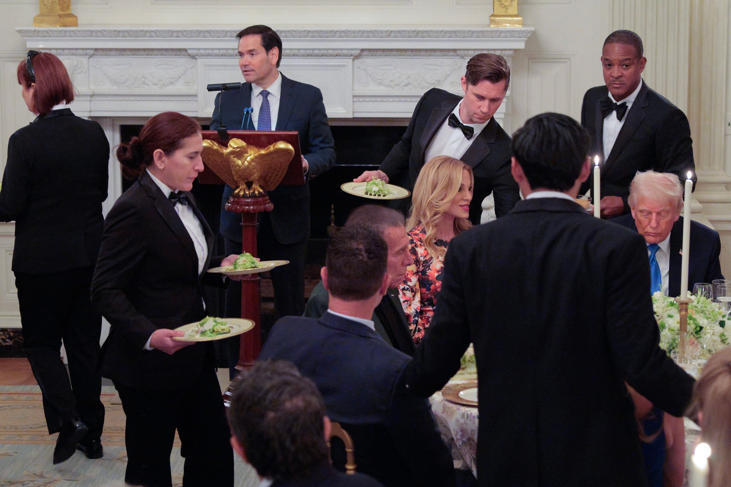 Waitstaff serve salads to seated guests at a formal dinner. A man stands at a podium speaking, while others are engaged in conversation at round tables in an elegant, ornate room.
