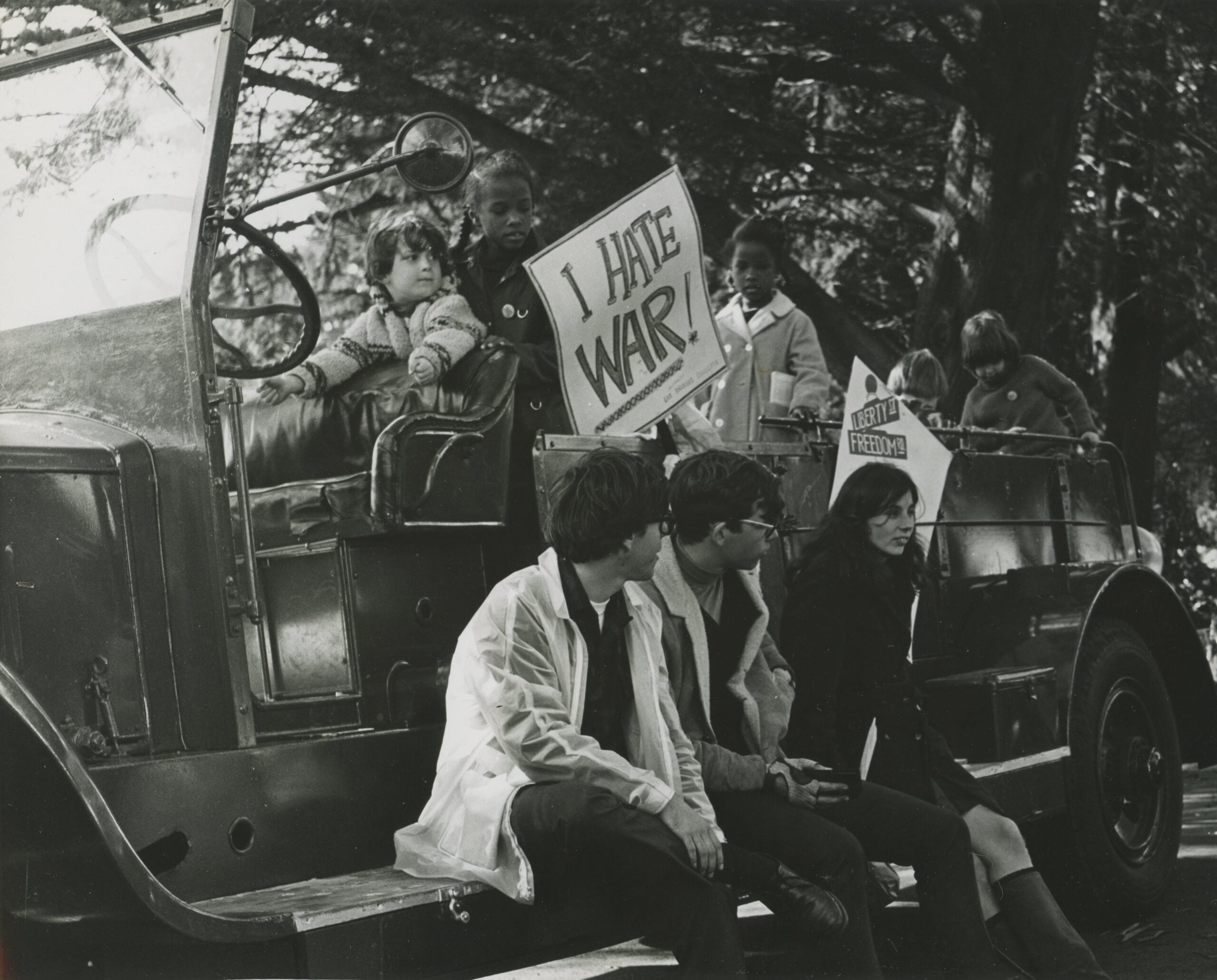 Several children sit on an old vehicle, one holding a sign reading “I HATE WAR!” Three adults and a child sit in front, with another sign that says “FIGHT FOR FREEDOM.” Trees are visible in the background.