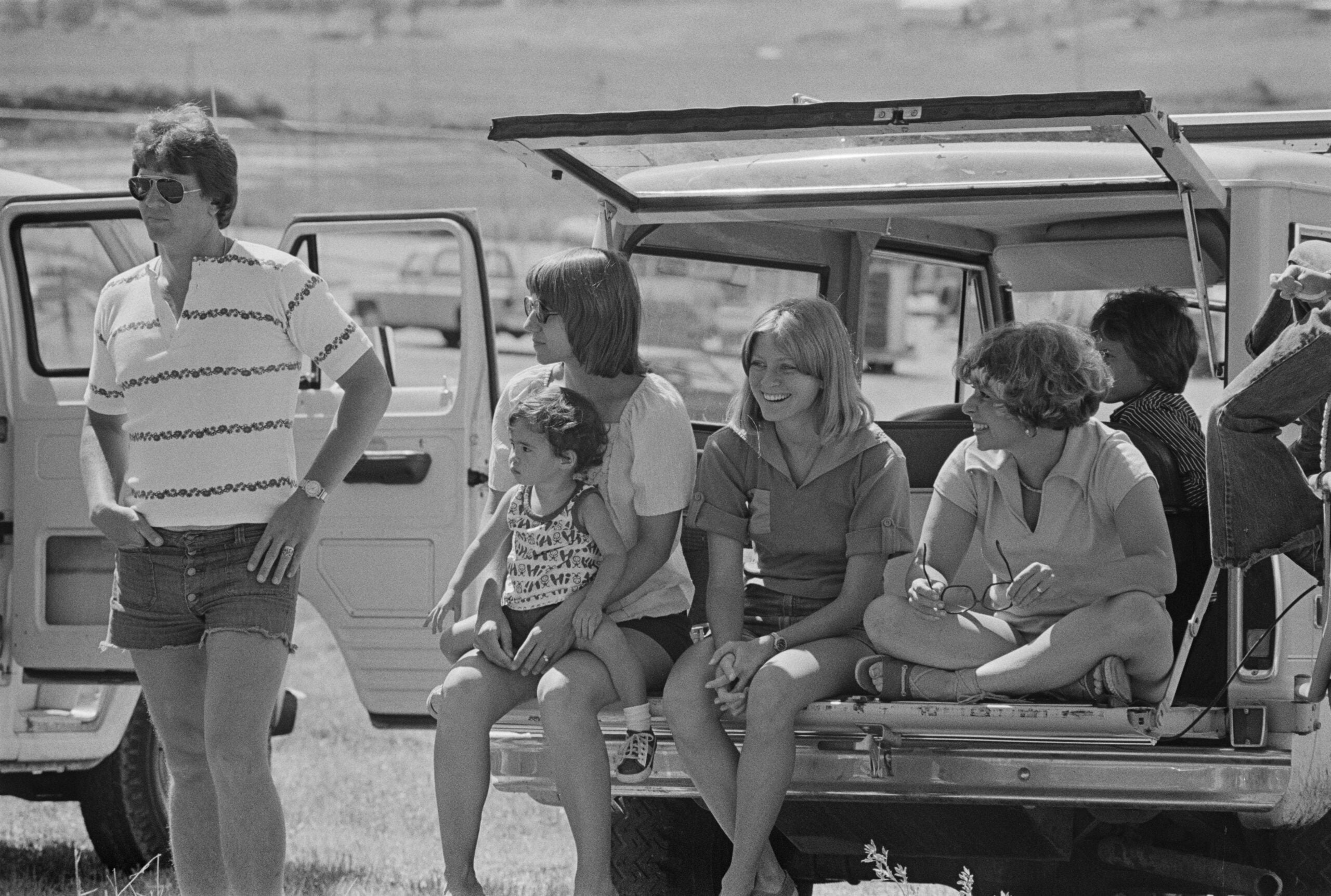 Six people, including children and adults, sit and stand near an open vehicle tailgate in a grassy outdoor setting, appearing relaxed and smiling on a sunny day.