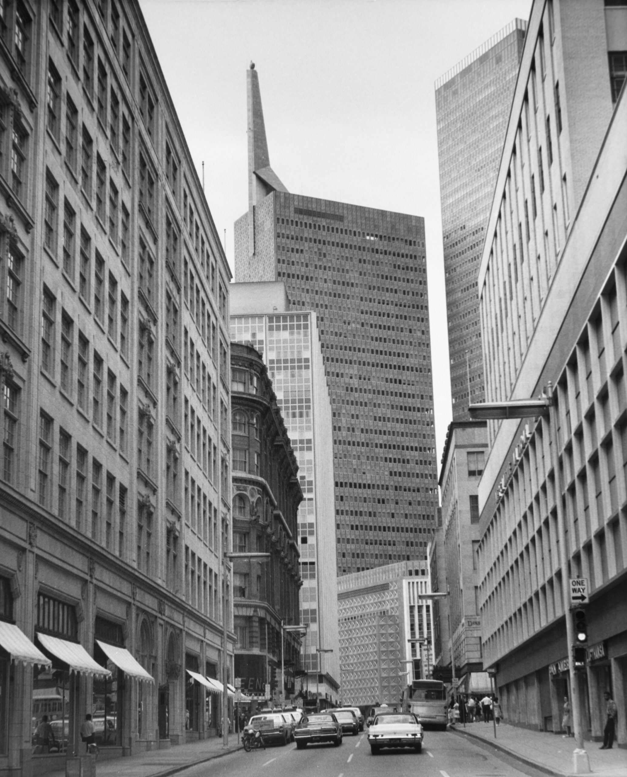 Black and white photo of a city street lined with tall buildings. Cars are parked along the curb, and a few are driving down the road. The Transamerica Pyramid is visible in the background.