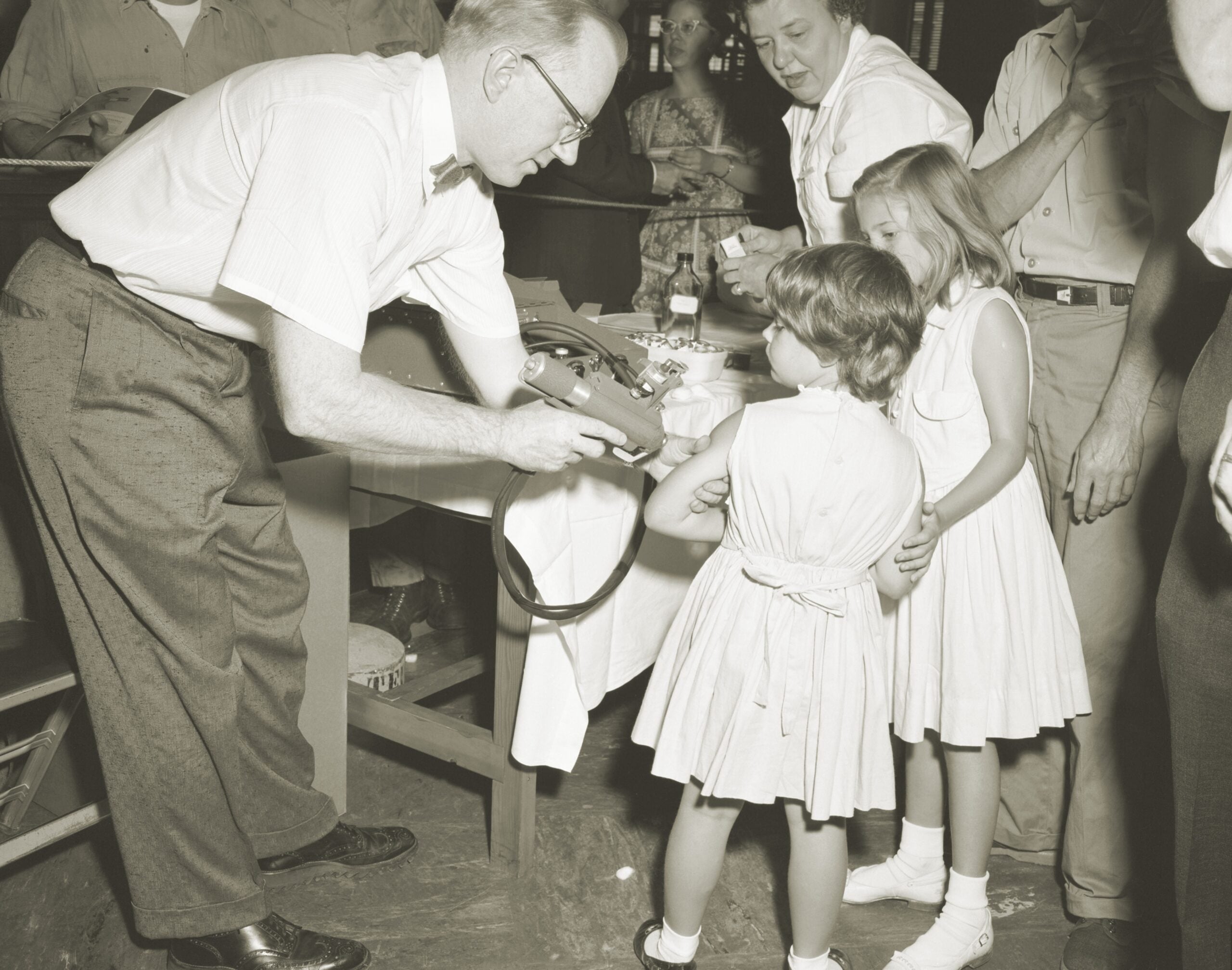 A man in glasses shows a camera to two young girls in dresses at an indoor gathering. Several adults watch and smile in the background. The scene appears friendly and informal.