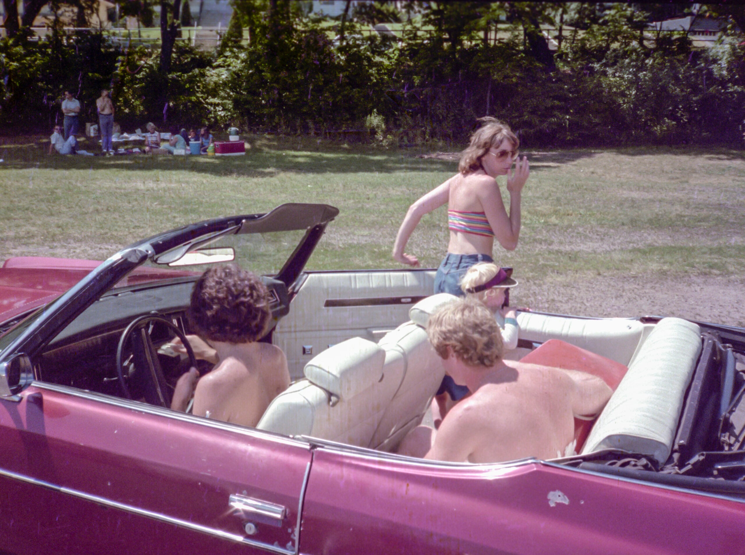 Three people sit in a red convertible with the top down, while a woman in a striped top stands outside the car, looking back and holding something near her face. There is a group sitting on the grass in the background.