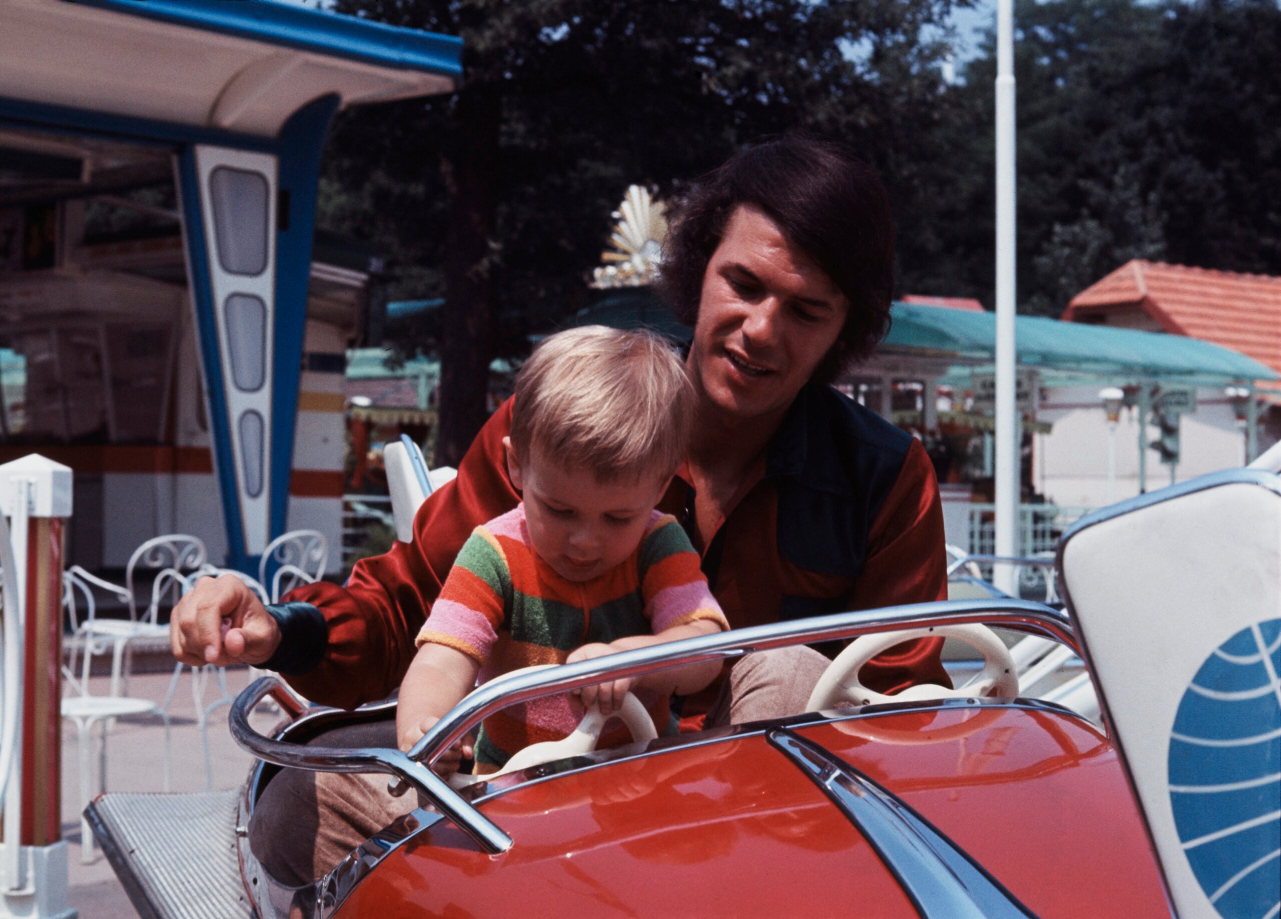 A man helps a small child sit in a red amusement park ride shaped like a car. The child wears a striped sweater and is focused on the steering wheel. Outdoor seating and greenery are visible in the background.