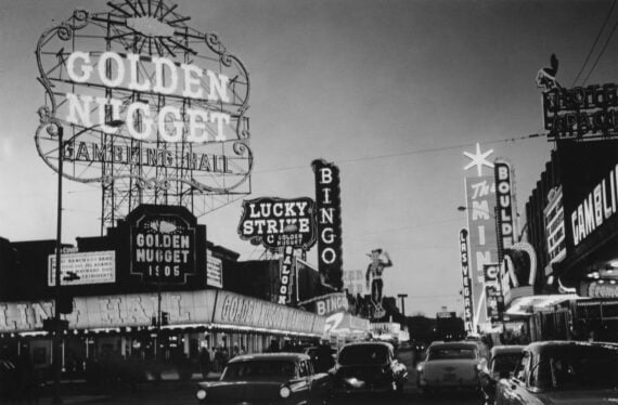 Black and white photo of a busy street in Las Vegas at night, featuring neon signs for places like the Golden Nugget, Lucky Strike, Bingo, and other casinos, with vintage cars on the road.