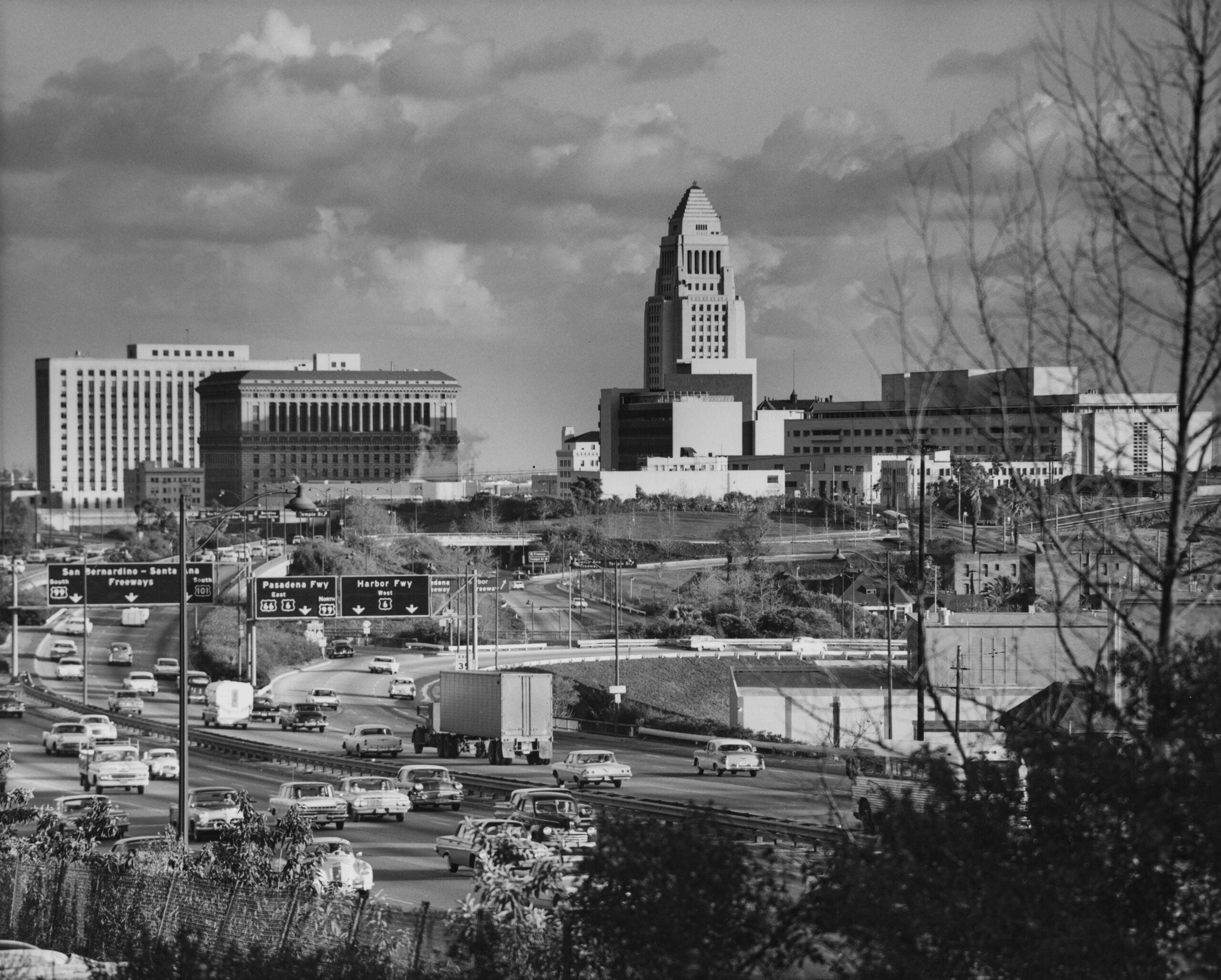 Major American cities 50 years ago. Black-and-white photo of a busy freeway with cars and trucks, leading toward tall government buildings and a distinctive tower in the background, under a partly cloudy sky.
