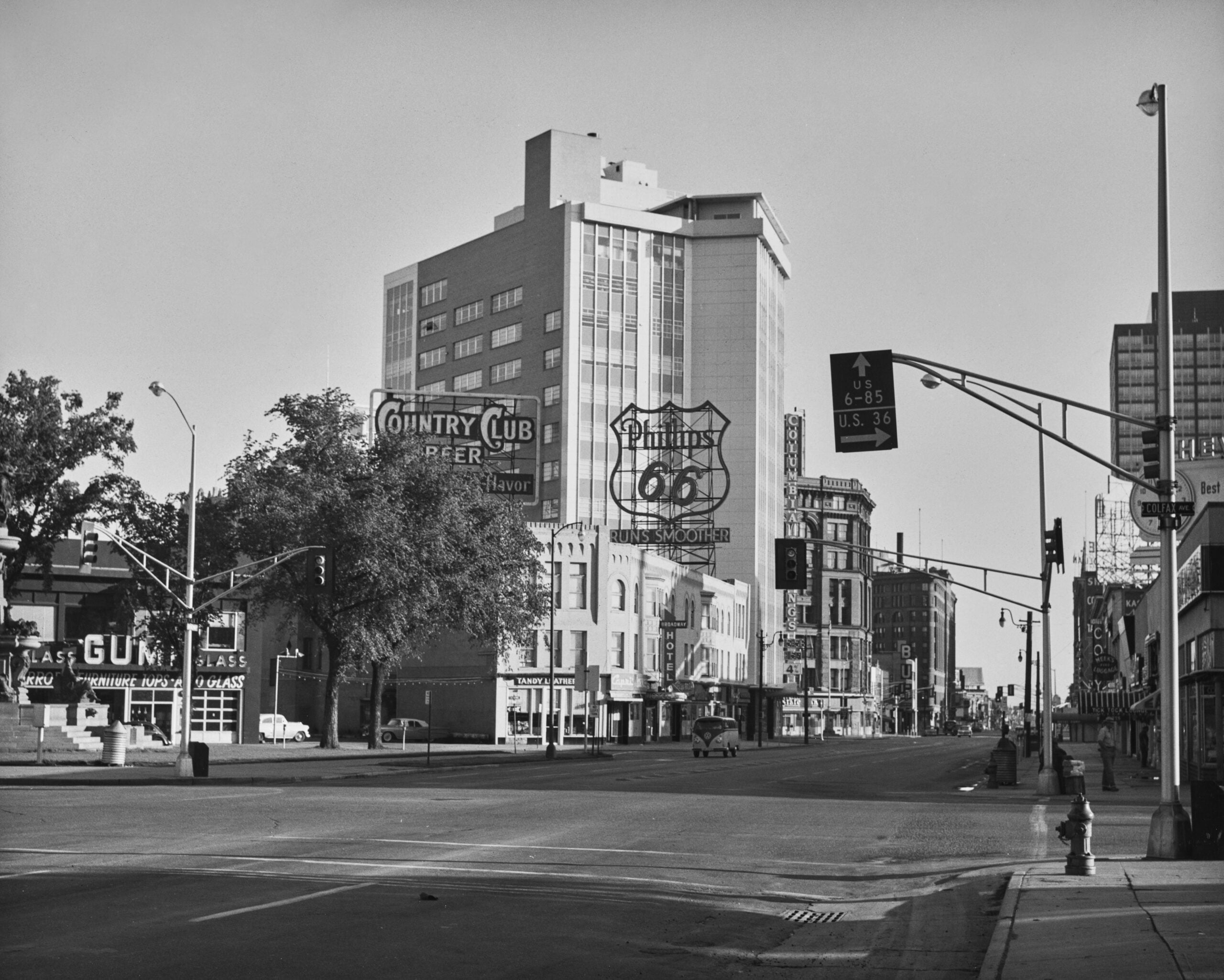 A black and white photo of a quiet downtown street with tall buildings, vintage signs for Route 66, Country Club Beer, and Phillips 66, plus traffic lights and minimal cars or people.