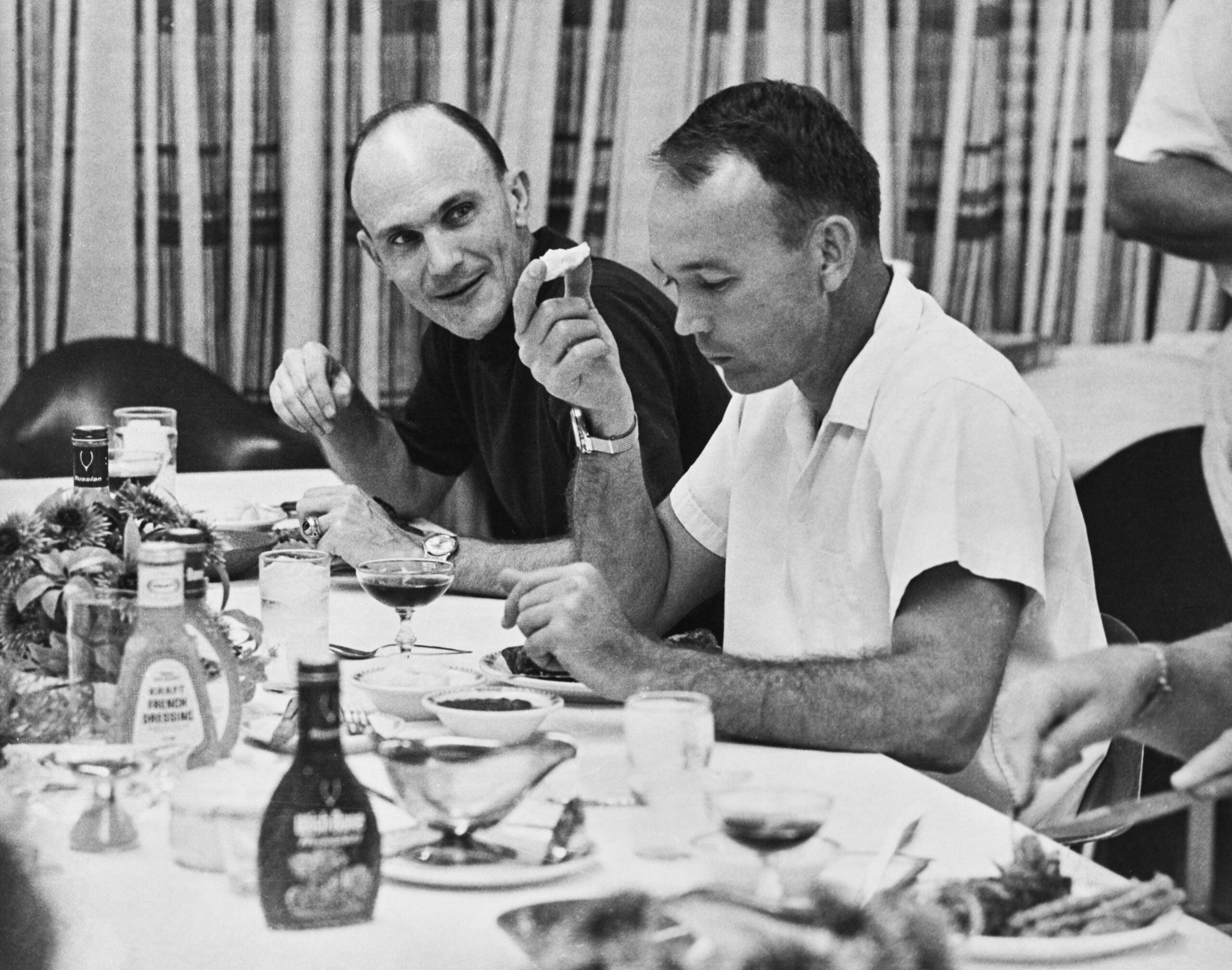 Two men sit at a dinner table, one eating while the other looks at him and smiles. Bottles of salad dressing and various dishes are visible on the table. The background shows patterned curtains.