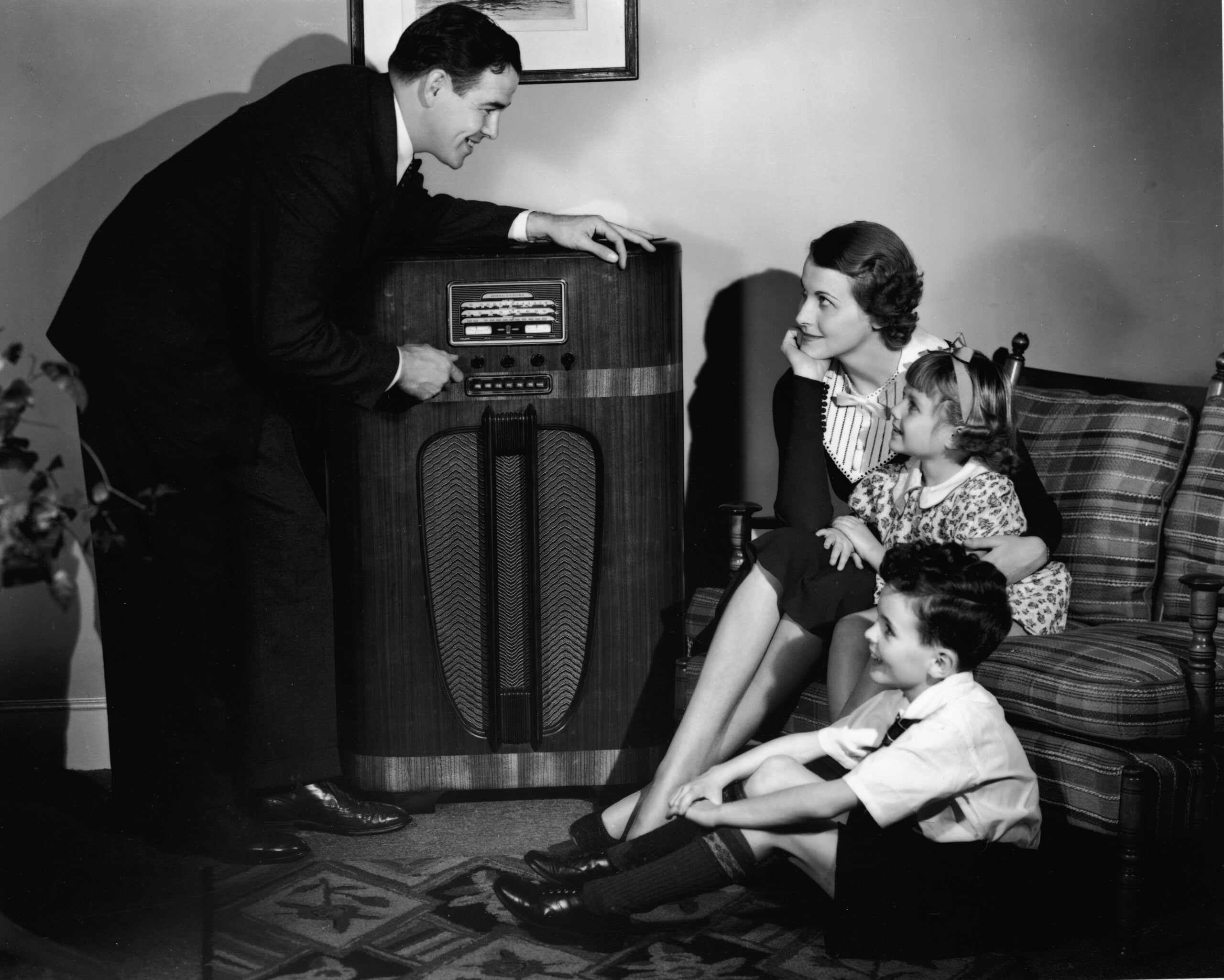 A man tunes an old-fashioned radio while a woman and two young children sit nearby on a couch, listening and smiling in a vintage living room setting.