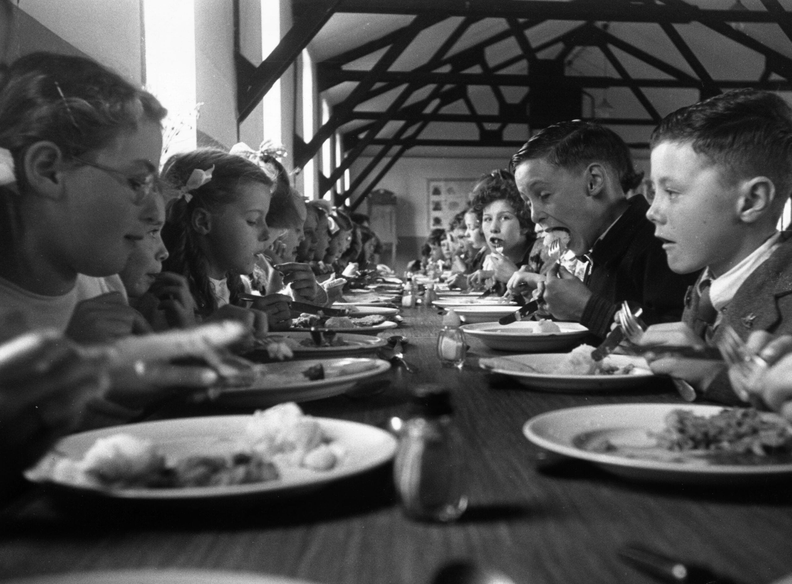 A group of children sit at a long table in a cafeteria, eating and laughing together. The room has high ceilings with exposed beams and sunlight streams through the windows.