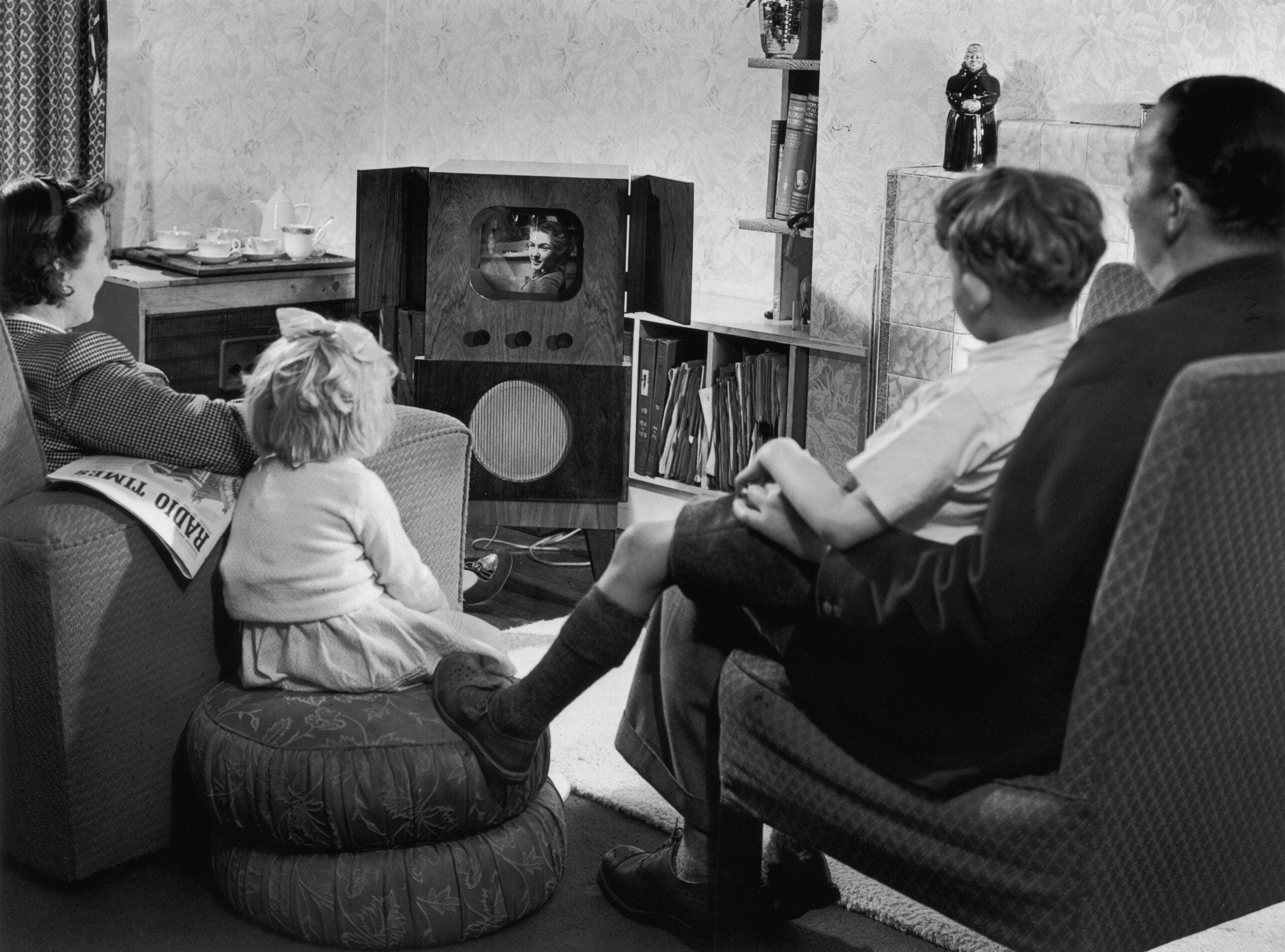 A family of four sits in a living room watching a small vintage television. The parents sit in chairs while two children sit on a cushioned ottoman. The room has patterned wallpaper and shelves with books.