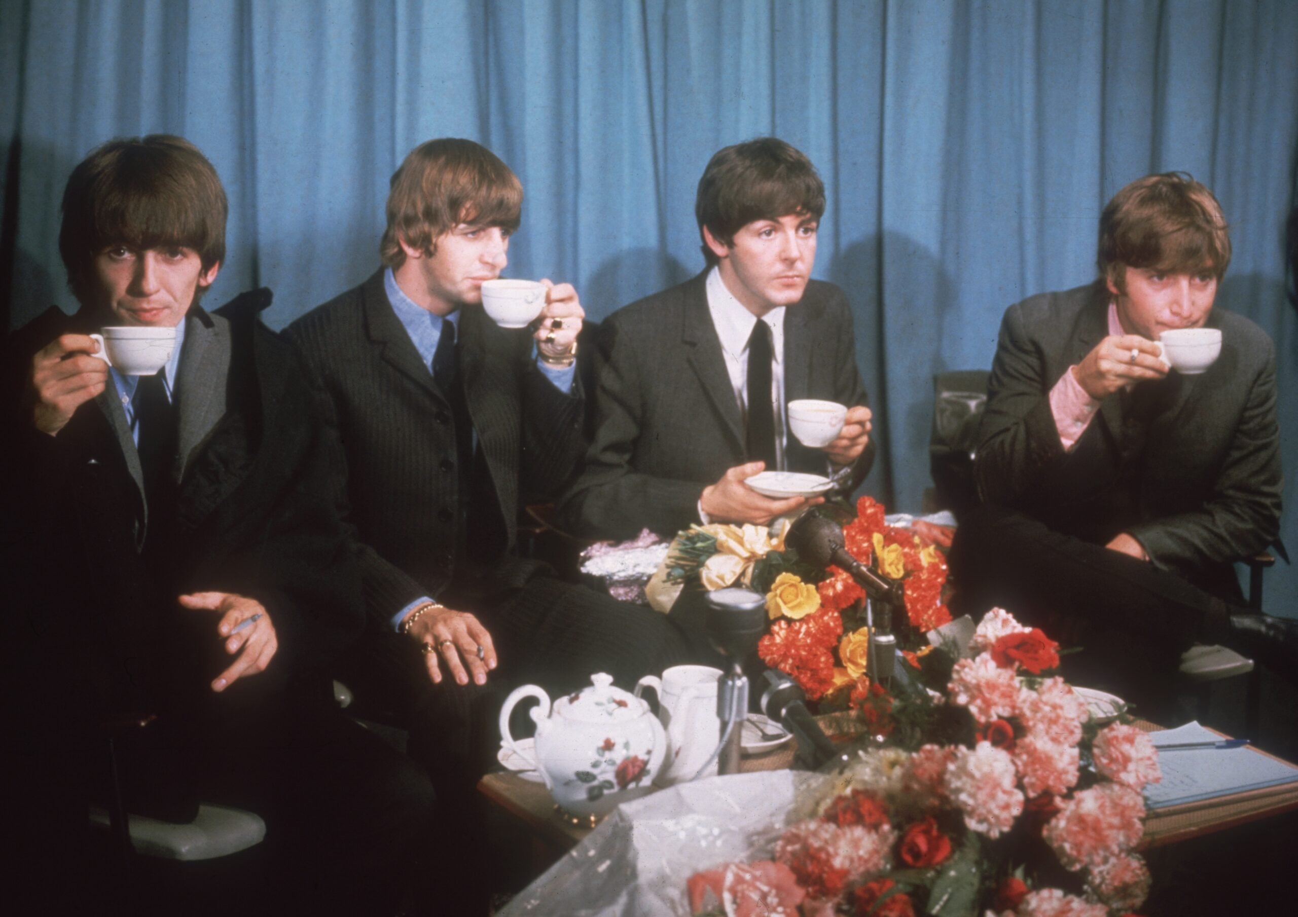 Four men in suits sit side by side, sipping tea from white cups in front of a blue curtain, with a table of flowers and a teapot in the foreground.