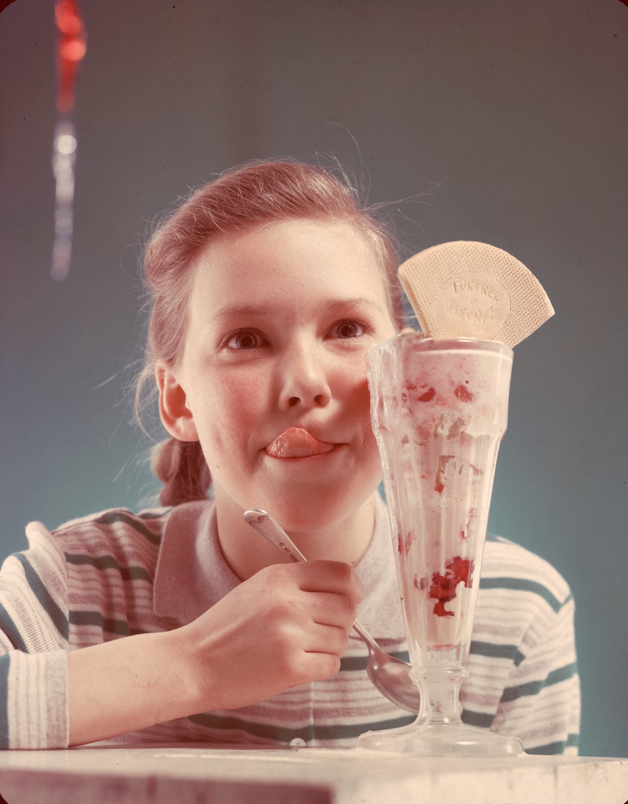 A young girl in a striped shirt playfully sticks out her tongue while sitting behind a tall glass of ice cream topped with a wafer. She holds a spoon and smiles, with a blue background behind her.