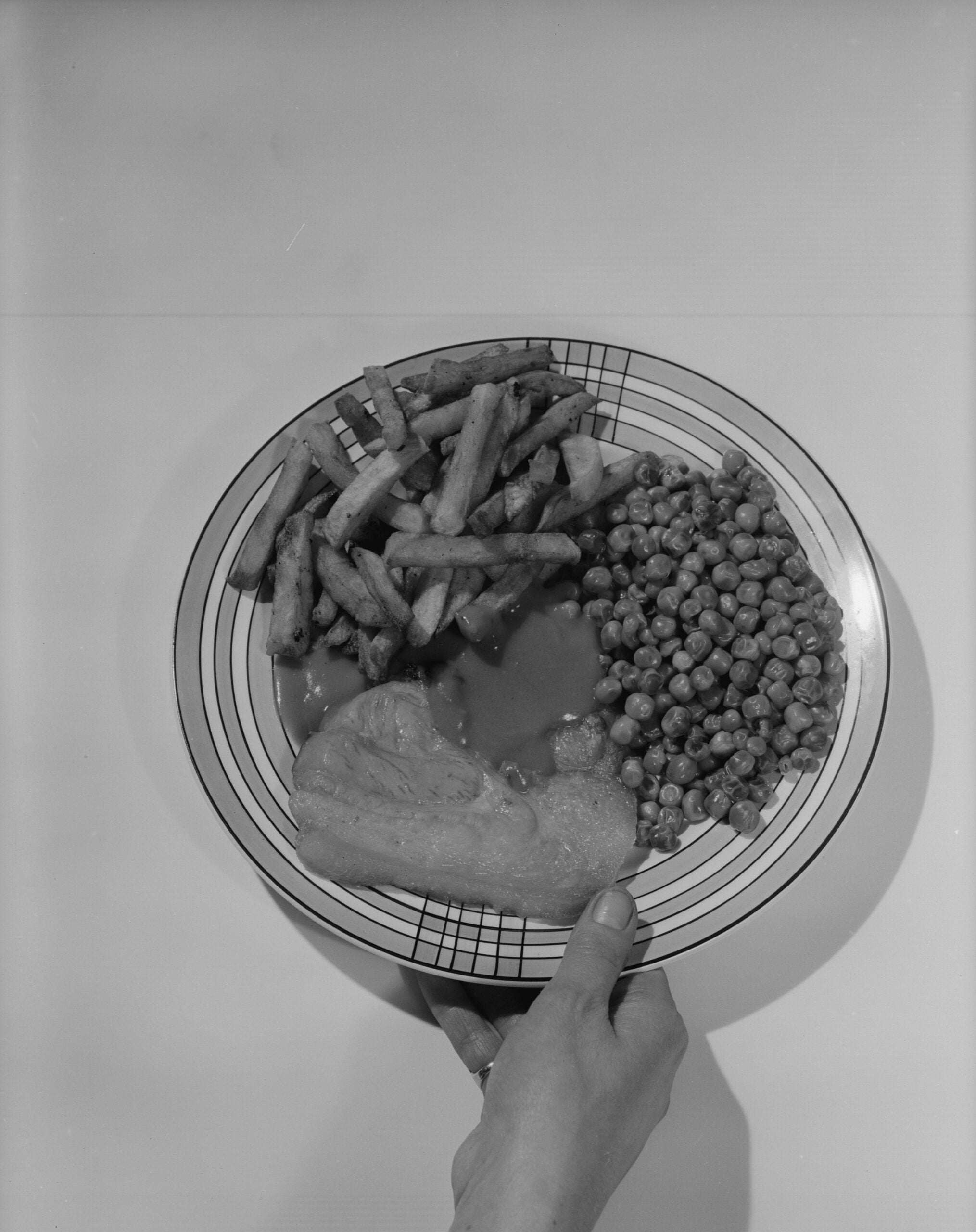 A hand holds a plate with French fries, green peas, and a piece of meat covered in gravy, shot in black and white.