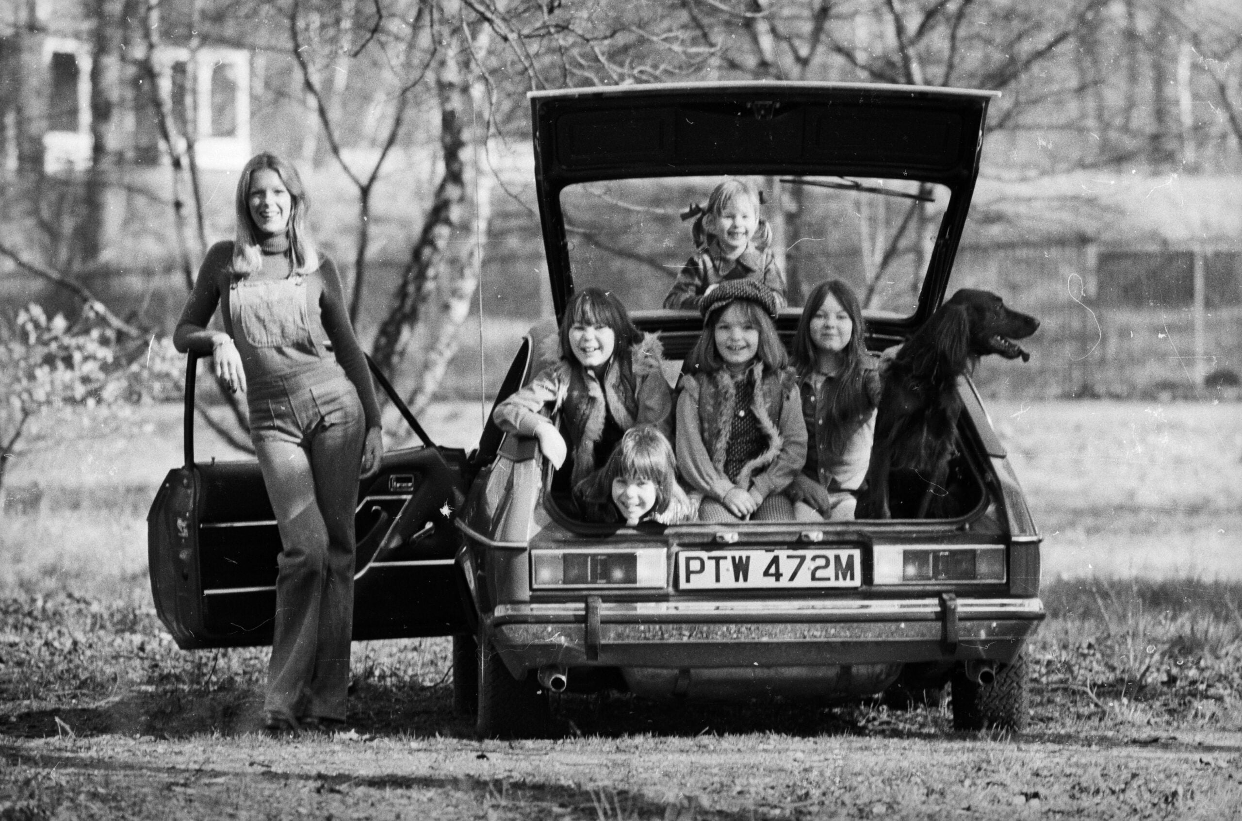 A woman in overalls stands by a car with its trunk open, while five children and a dog sit or lean inside the trunk. Trees and a blurred house are visible in the background.