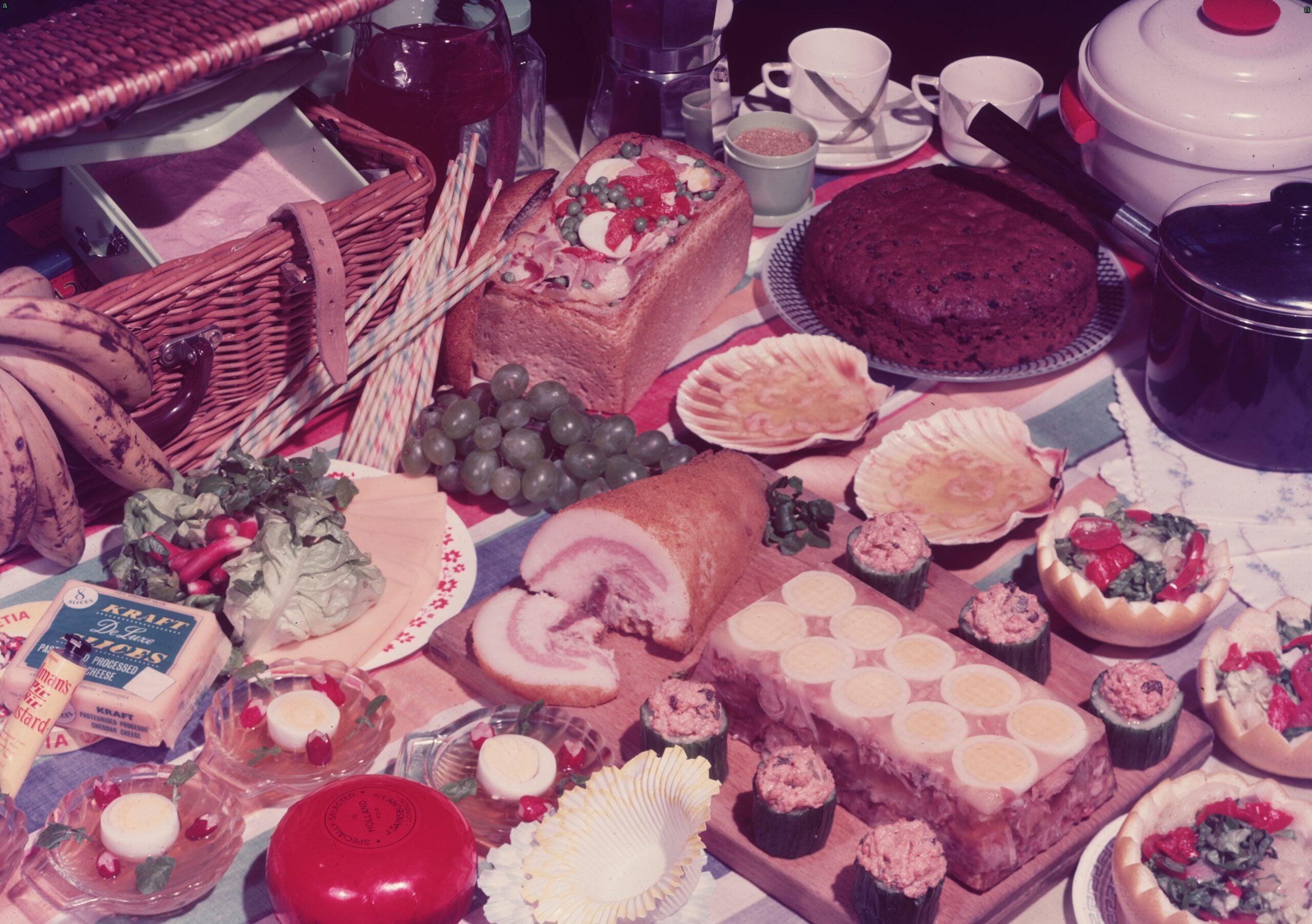 A picnic spread with fruits, bread, cheese, cake, salad, butter, cupcakes, and various dishes arranged on a table with cups, plates, and a picnic basket.
