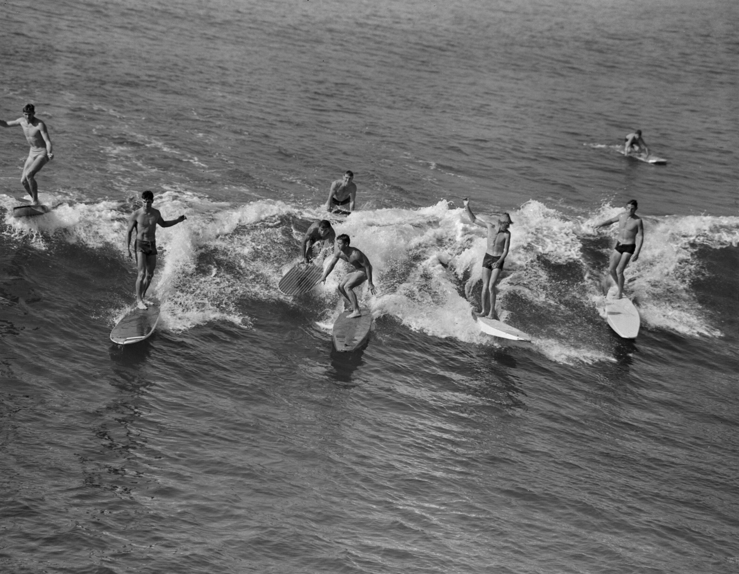Six surfers ride a small wave together on longboards, close to each other, while another person paddles nearby. The image is in black and white, capturing a lively moment on the water.