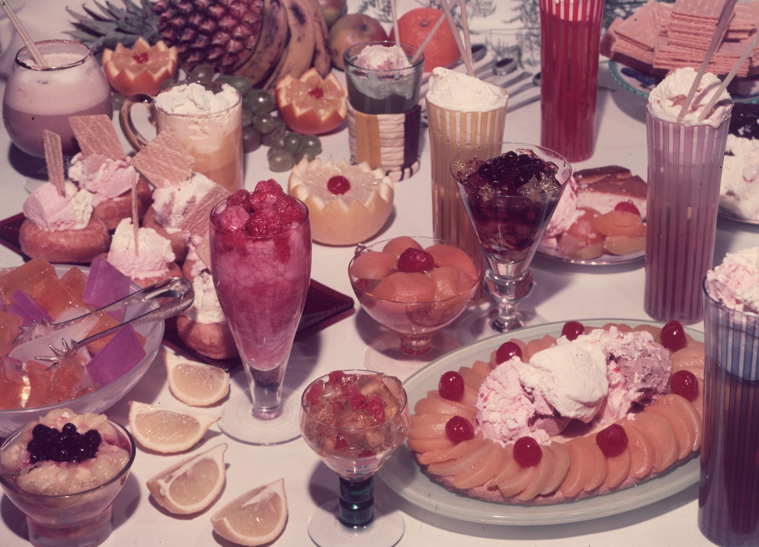 A table covered with various desserts including ice cream, parfaits, jellies, fruit salads, and drinks, surrounded by sliced lemons, fruit, and wafers.