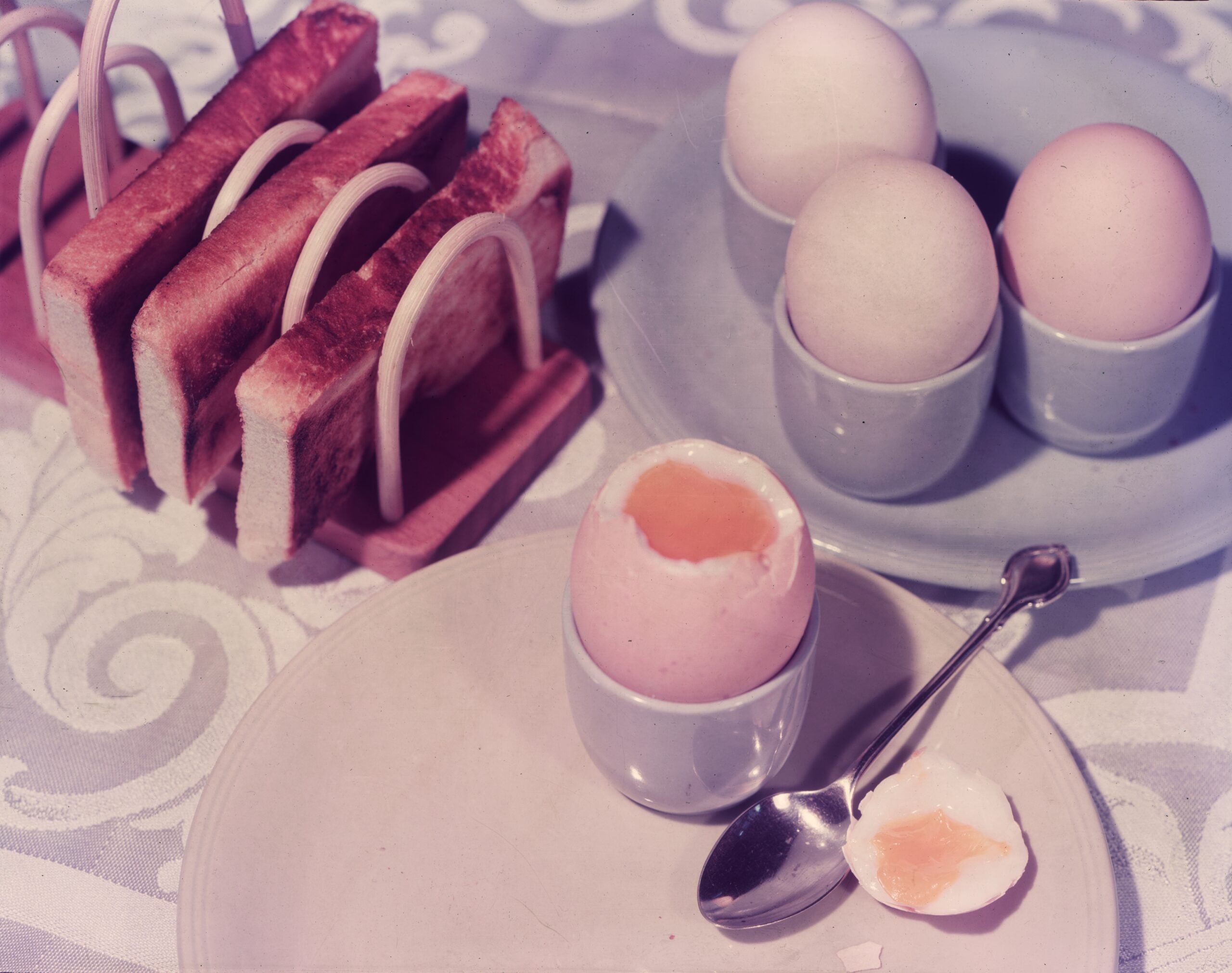 Meals from the 40s and 50s . Soft-boiled eggs in egg cups with a spoon, and slices of toast in a toast rack, are set on a patterned tablecloth.