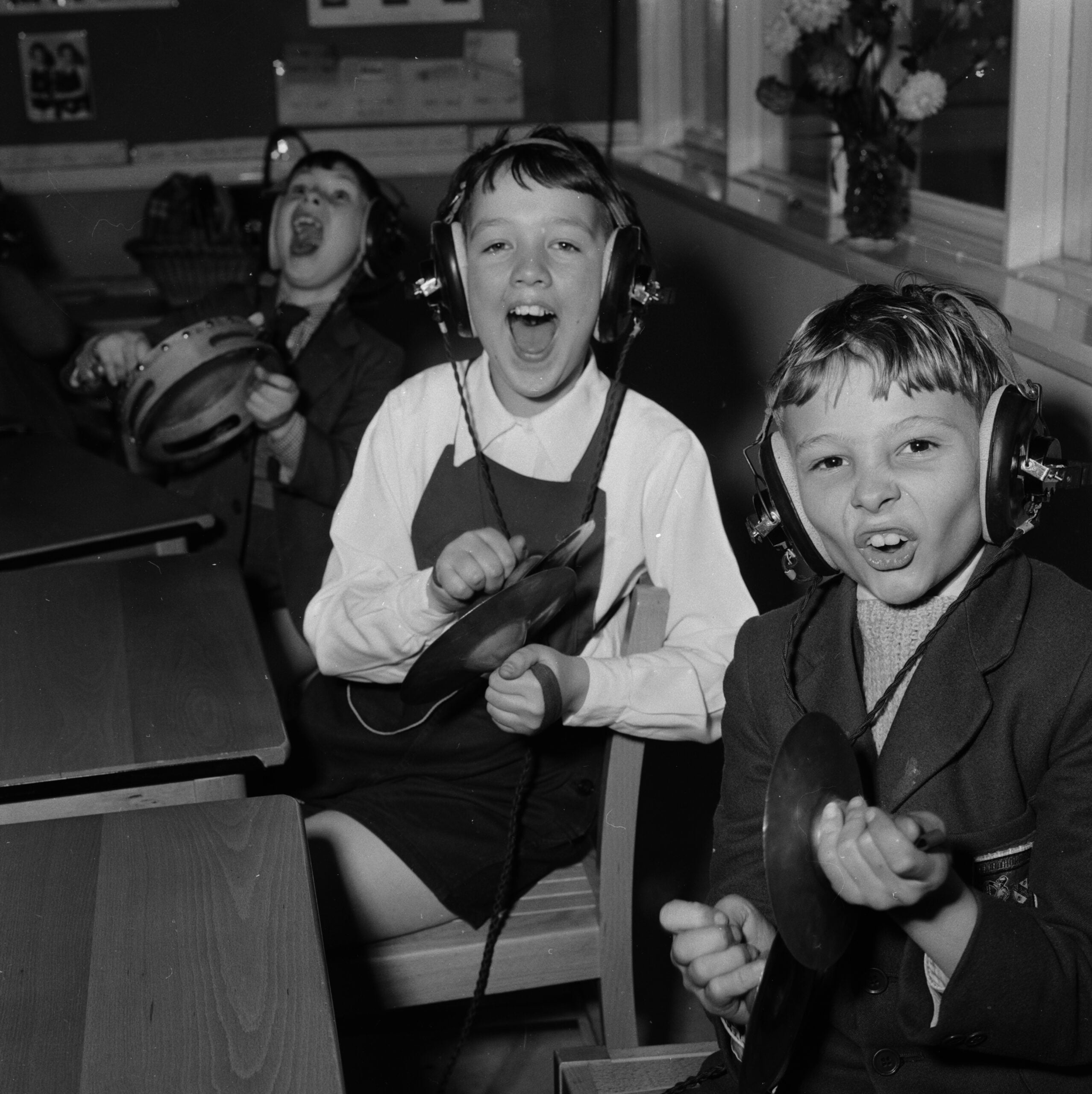 Three children, wearing headphones and school uniforms, enthusiastically play musical instruments and sing at their desks in a classroom. Two hold tambourines, and one holds a cymbal. All three look happy and energetic.