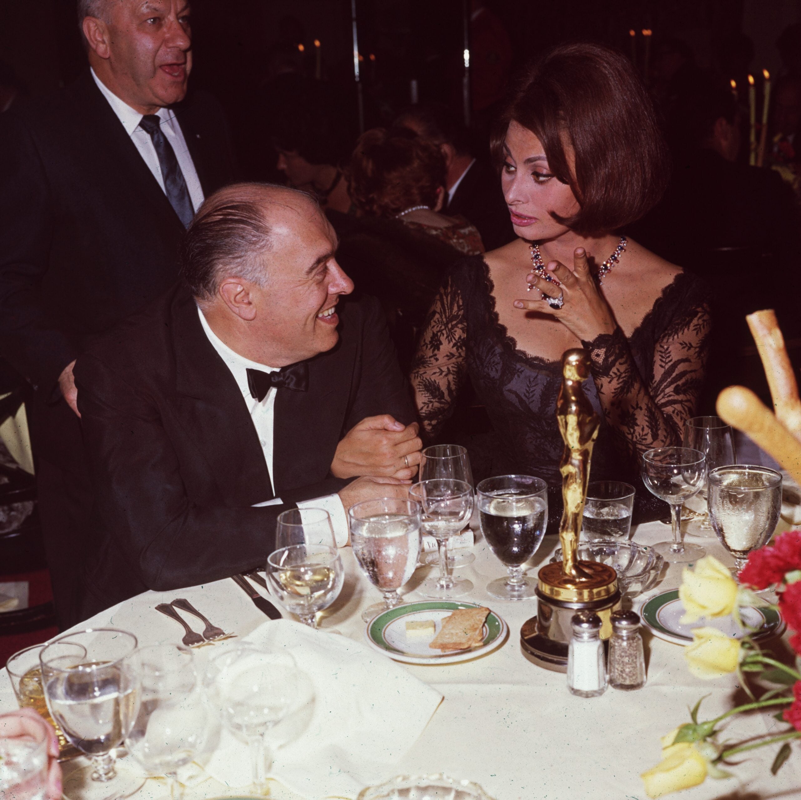 A woman in a black lace dress sits at a dinner table with a gold statue and a man in a tuxedo. They are smiling and talking at a formal event. Other guests are visible in the background.