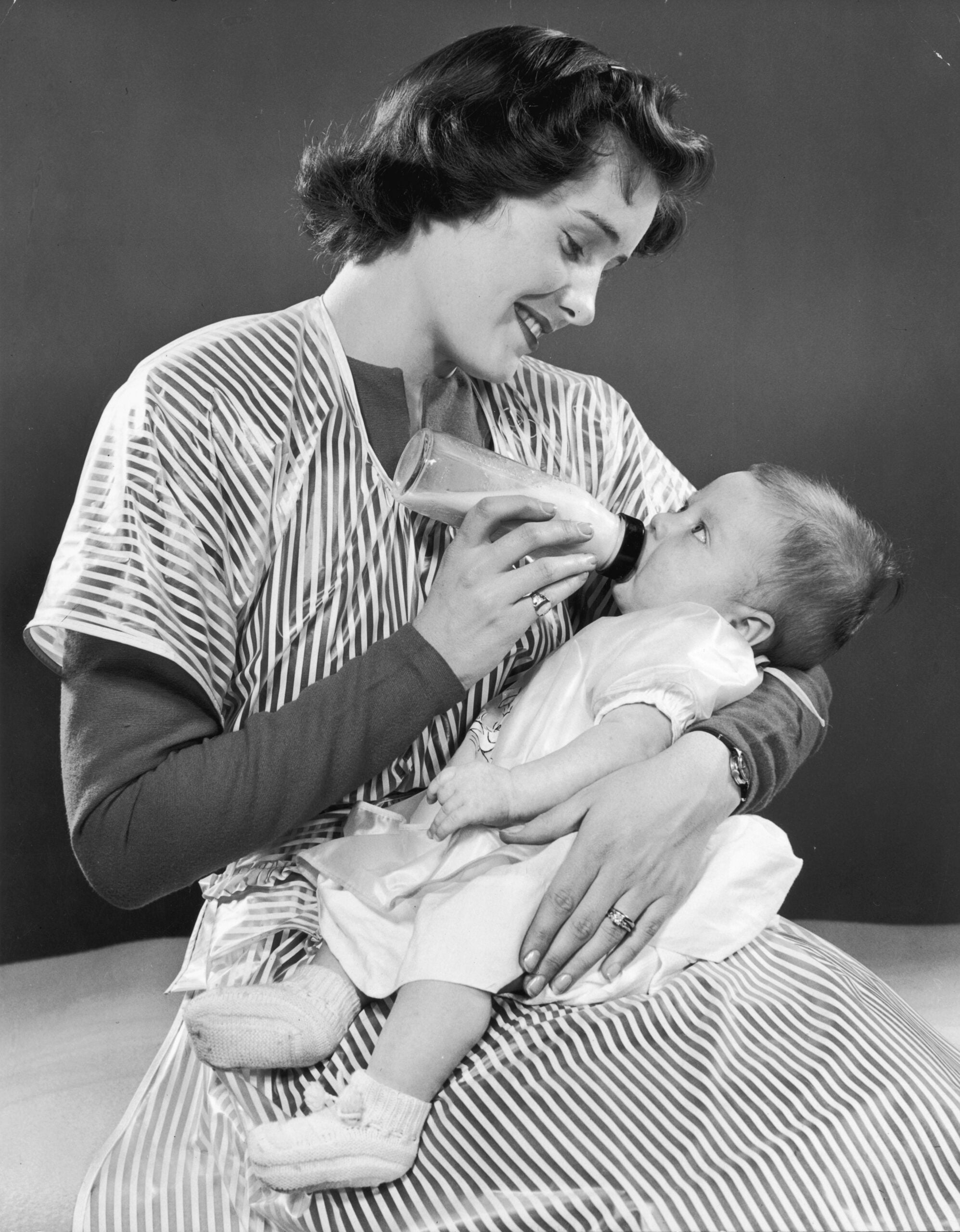 A woman in a striped dress smiles while bottle-feeding a baby dressed in white. The baby lies in her arms and looks up at her. The background is plain and dark.