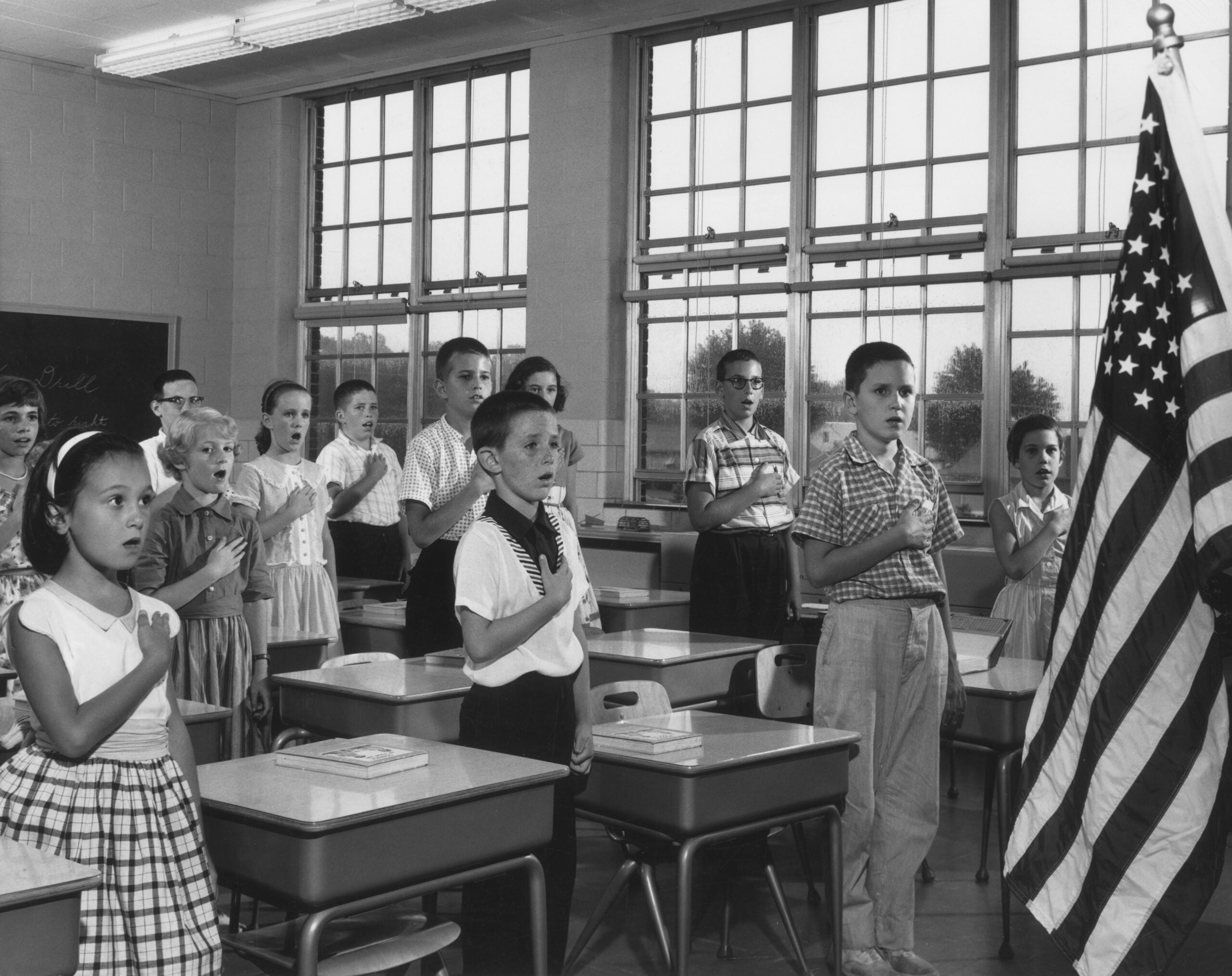 A group of children in a classroom stand with hands on their hearts, facing the American flag, reciting the Pledge of Allegiance. Desks and windows fill the background.