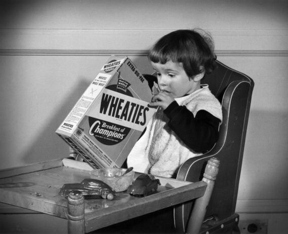 A young child sitting in a high chair looks at a large Wheaties cereal box, with a bowl and toy cars on the tray in front of them.