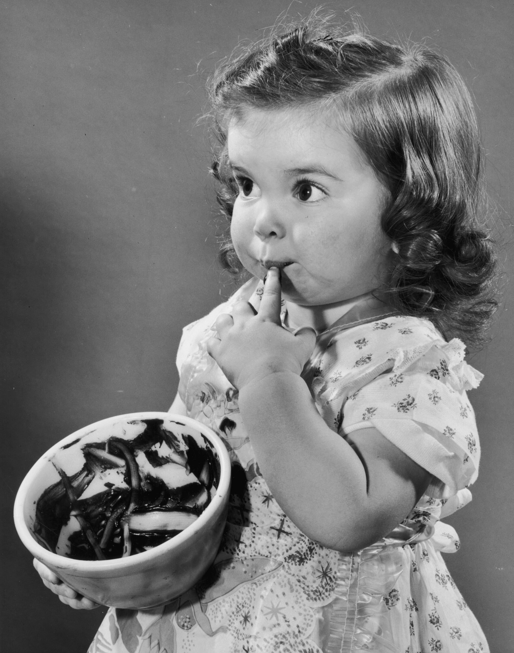 A young girl with curly hair licks chocolate off her finger while holding a bowl with chocolate remnants, wearing a light-patterned dress and looking slightly to the side.