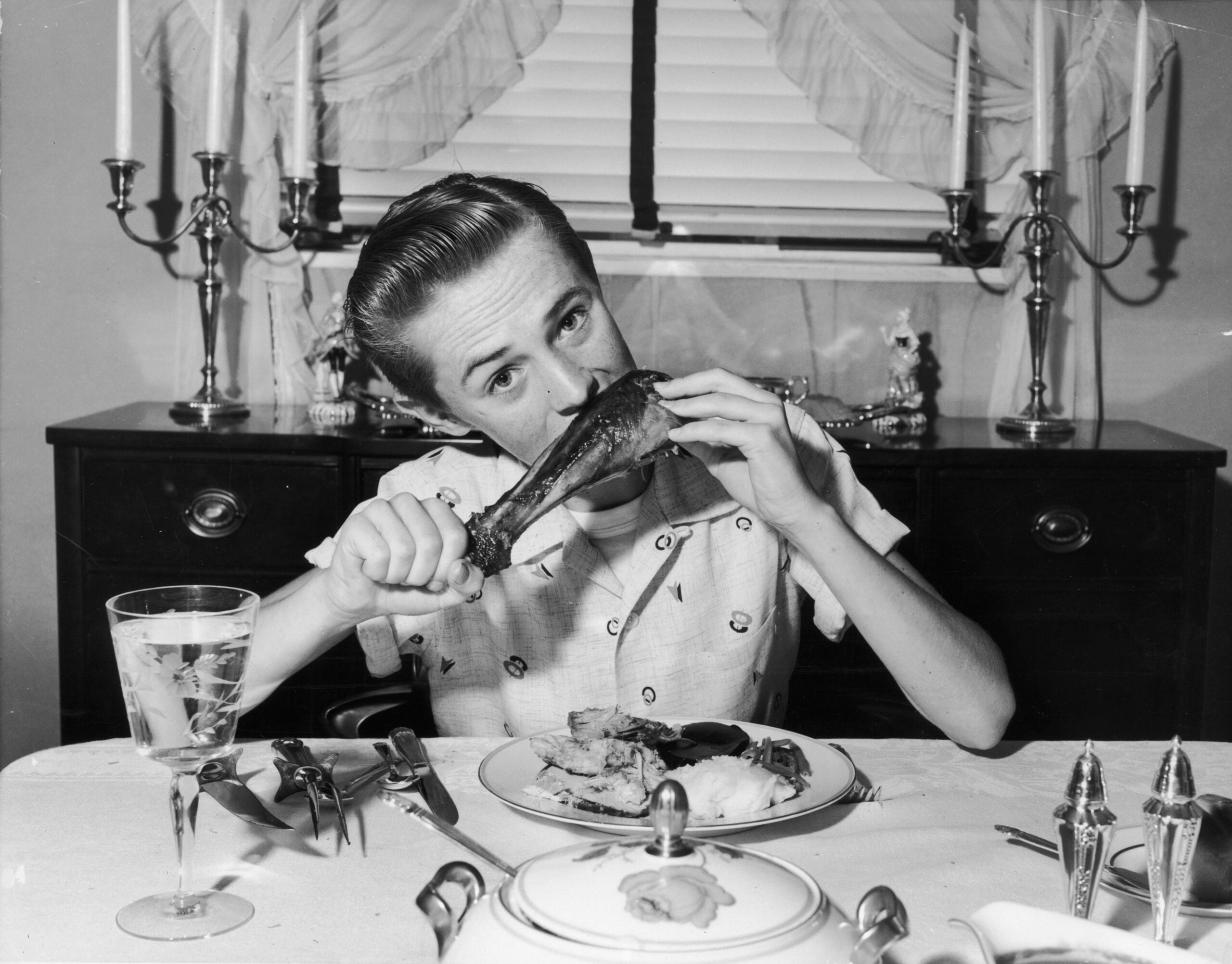 A young person sits at a dining table, biting into a large turkey leg. The table is set with food, silverware, and a glass of water. Candlesticks and a decorative cabinet are in the background.