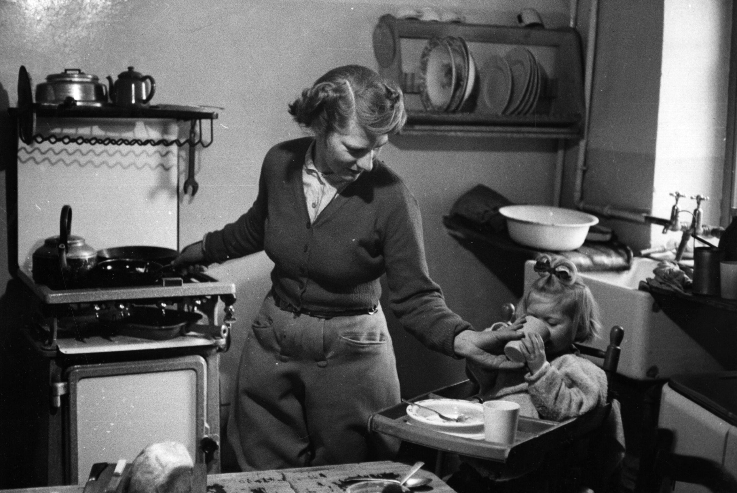 A woman cooks at a stove while feeding a young child in a high chair. The kitchen includes shelves with plates, a sink, and a cup and plate on the table. The scene appears to be from an earlier era.